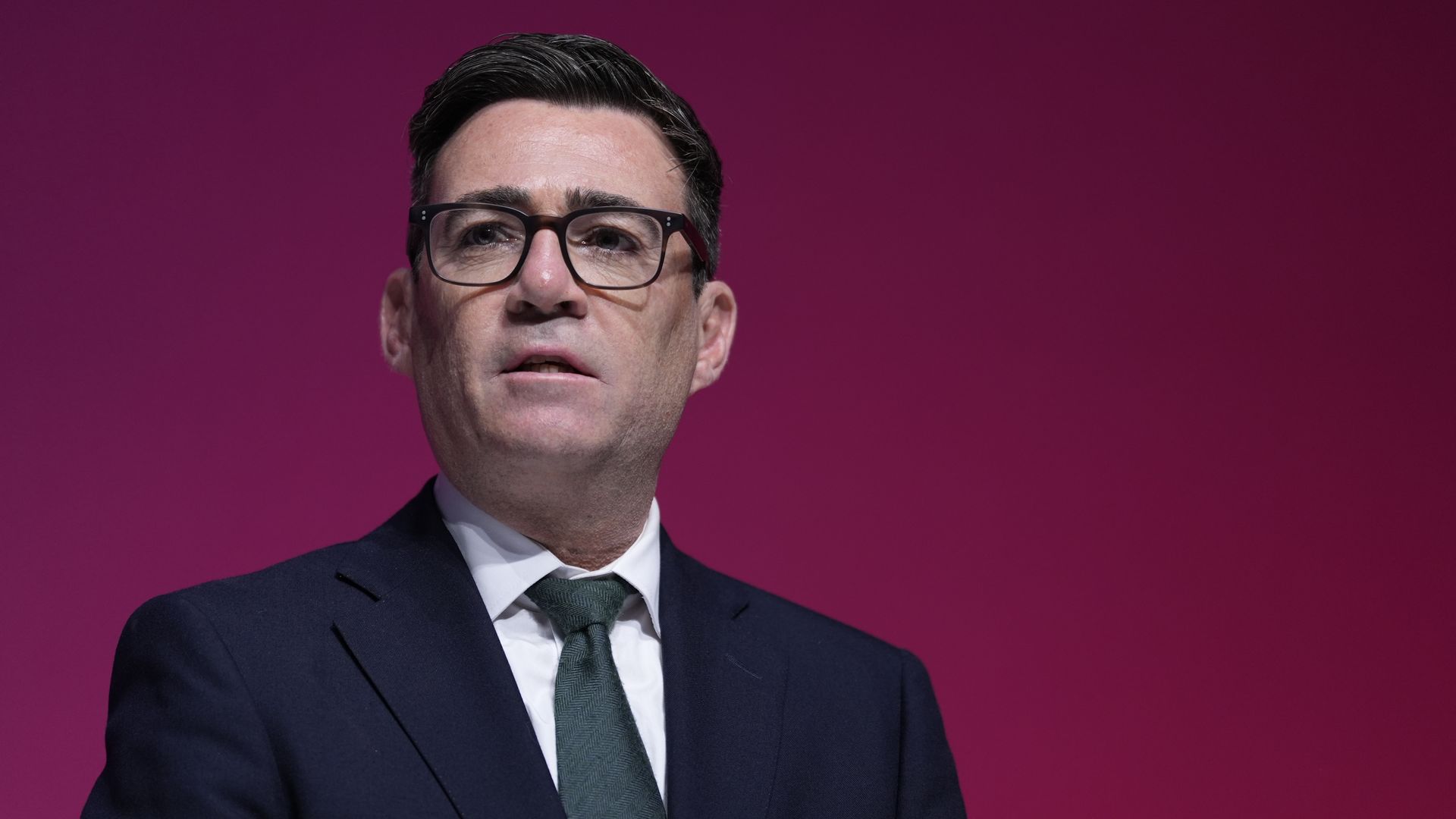 Portrait of a man, British Prime Minister Andy Burnham, with dark hair and glasses, wearing a dark suit, white shirt, and green tie, against a solid magenta background.