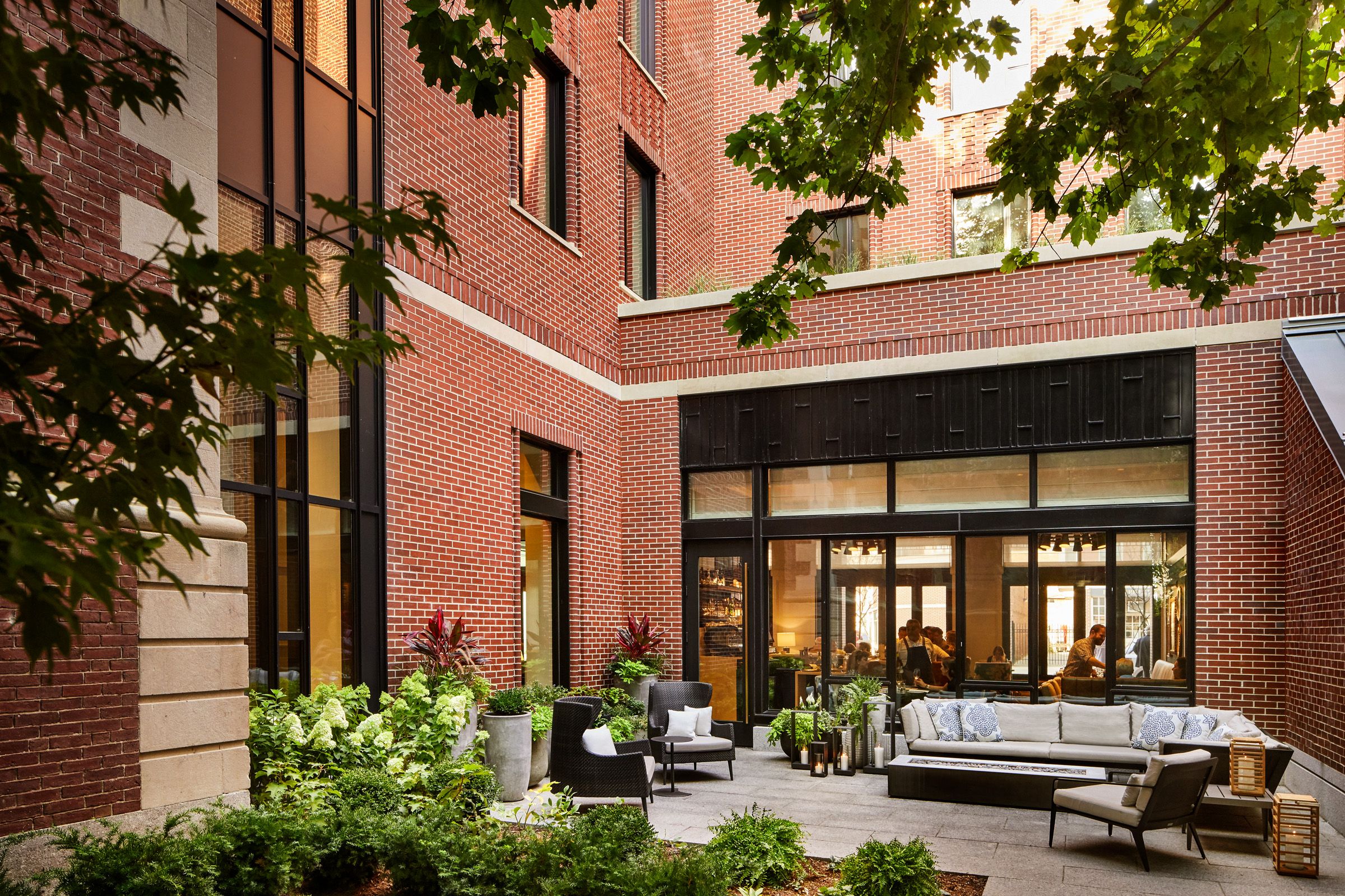 Red brick courtyard with black-framed glass doors and a modern outdoor seating area — gray sofa, black chairs and cushions. Potted plants line the patio; leafy trees overhead; people visible inside through windows.