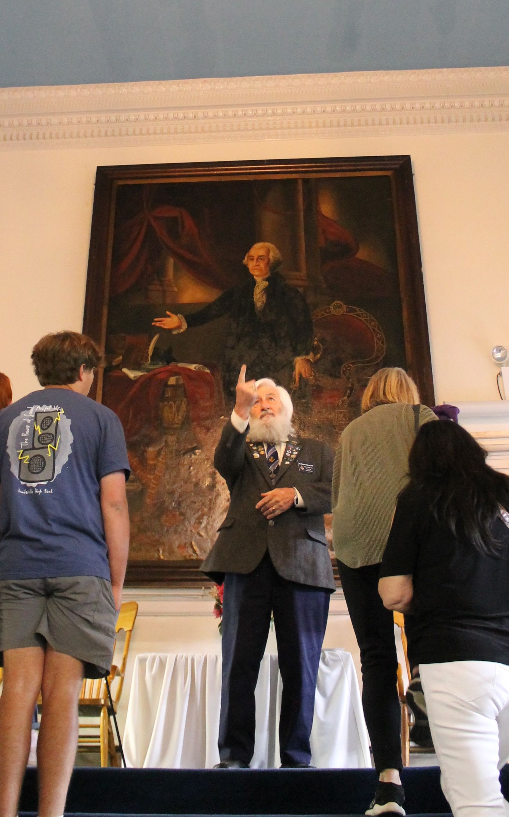An elderly man with a white beard and navy suit gestures upward in front of a large classical painting of a man in dark clothing with an outstretched arm. Several people stand nearby.