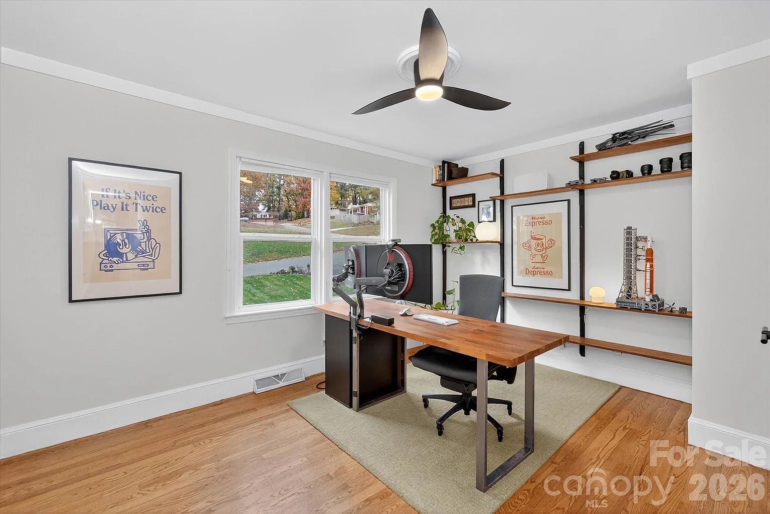 Modern home office with wood floor, light gray walls, wooden desk, black swivel chair, dual window, ceiling fan, framed posters reading "If It's Nice Play It Twice" and "More Espresso Less Depresso," wall shelves with decor.