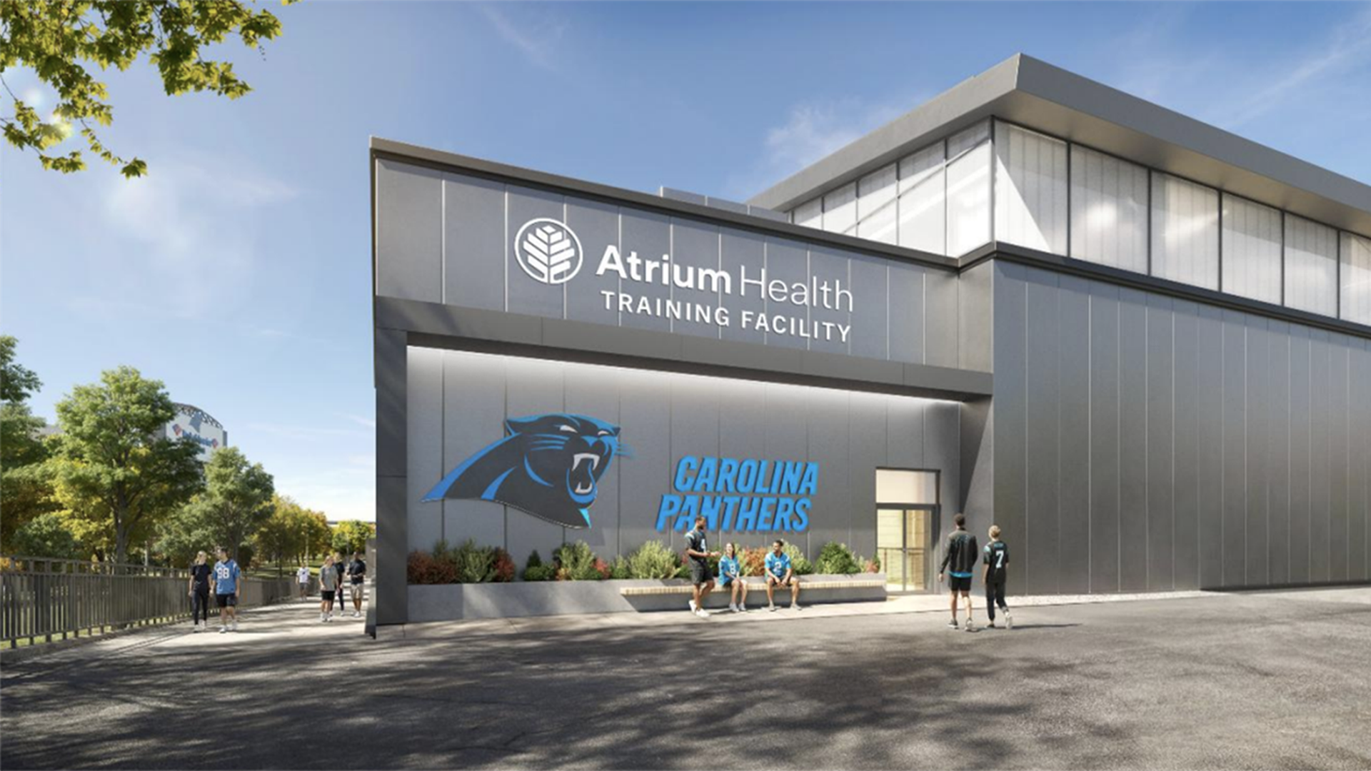 Exterior view of Atrium Health Training Facility with a Carolina Panthers logo and name on the wall; several people sit on a bench and walk outside on a sunny day.