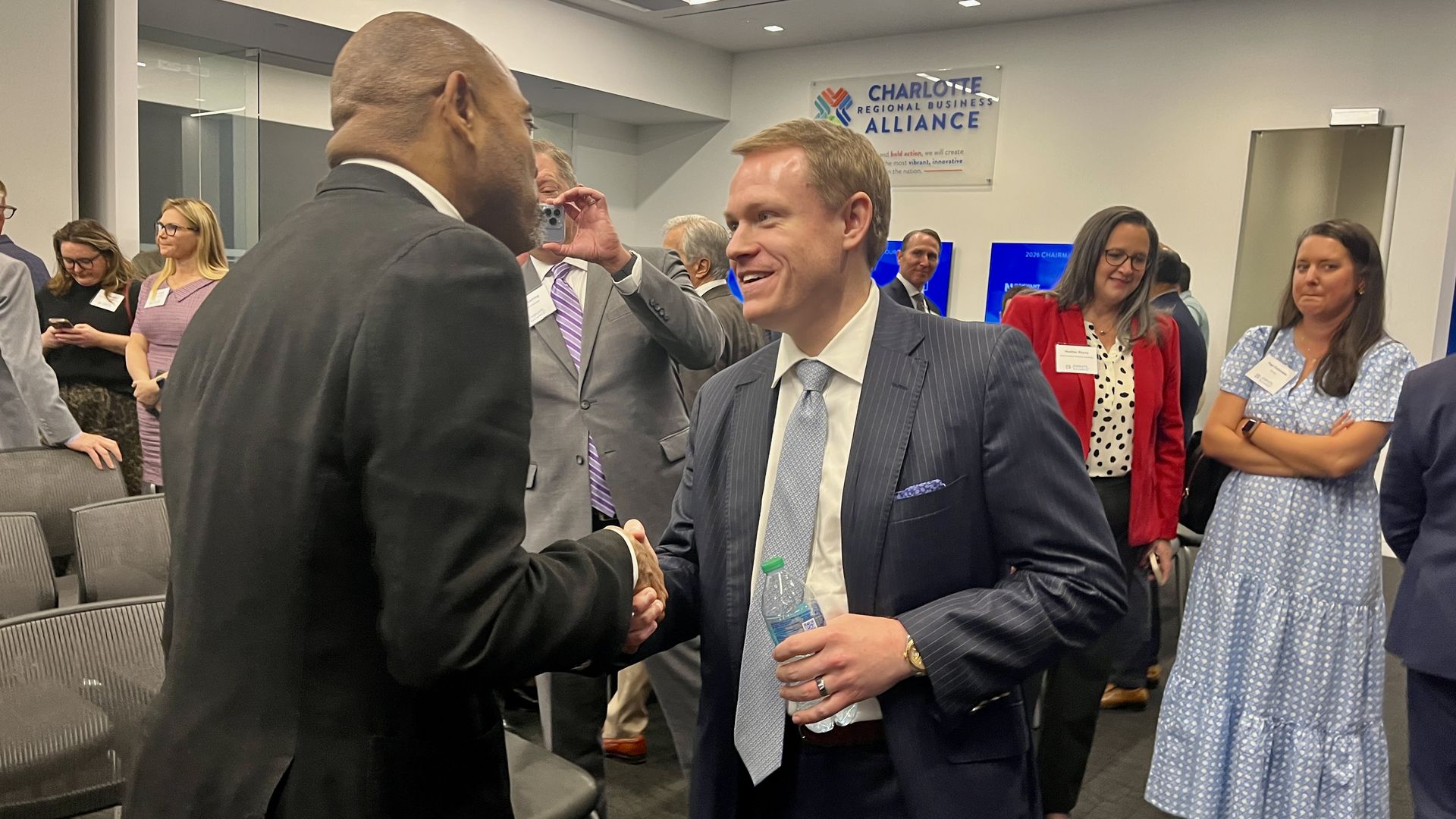 Two men in suits shake hands at a professional event, one holding a water bottle. In the background, attendees chat near a Charlotte Regional Business Alliance sign.