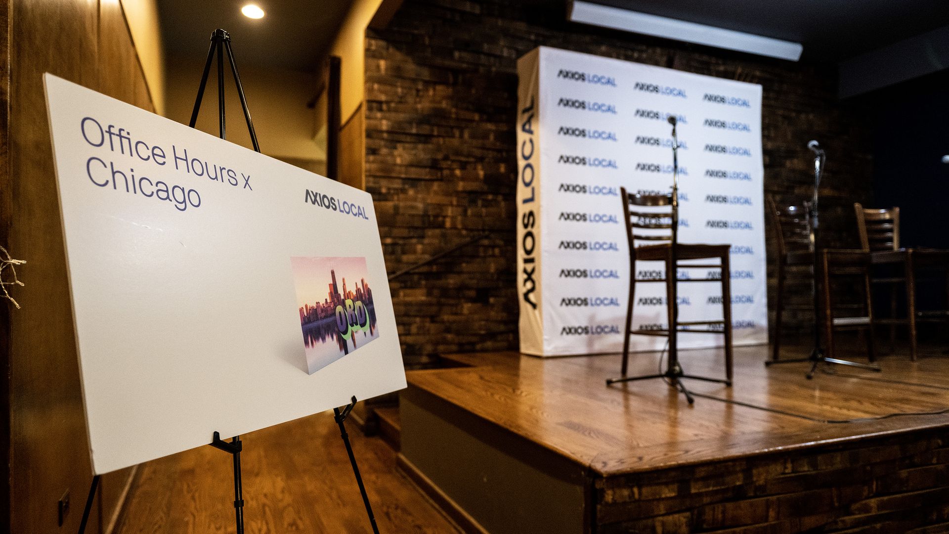Empty stage with wooden chairs and microphones, Axios Local backdrop, and a sign on a stand reading "Office Hours x Chicago" with a Chicago skyline image.