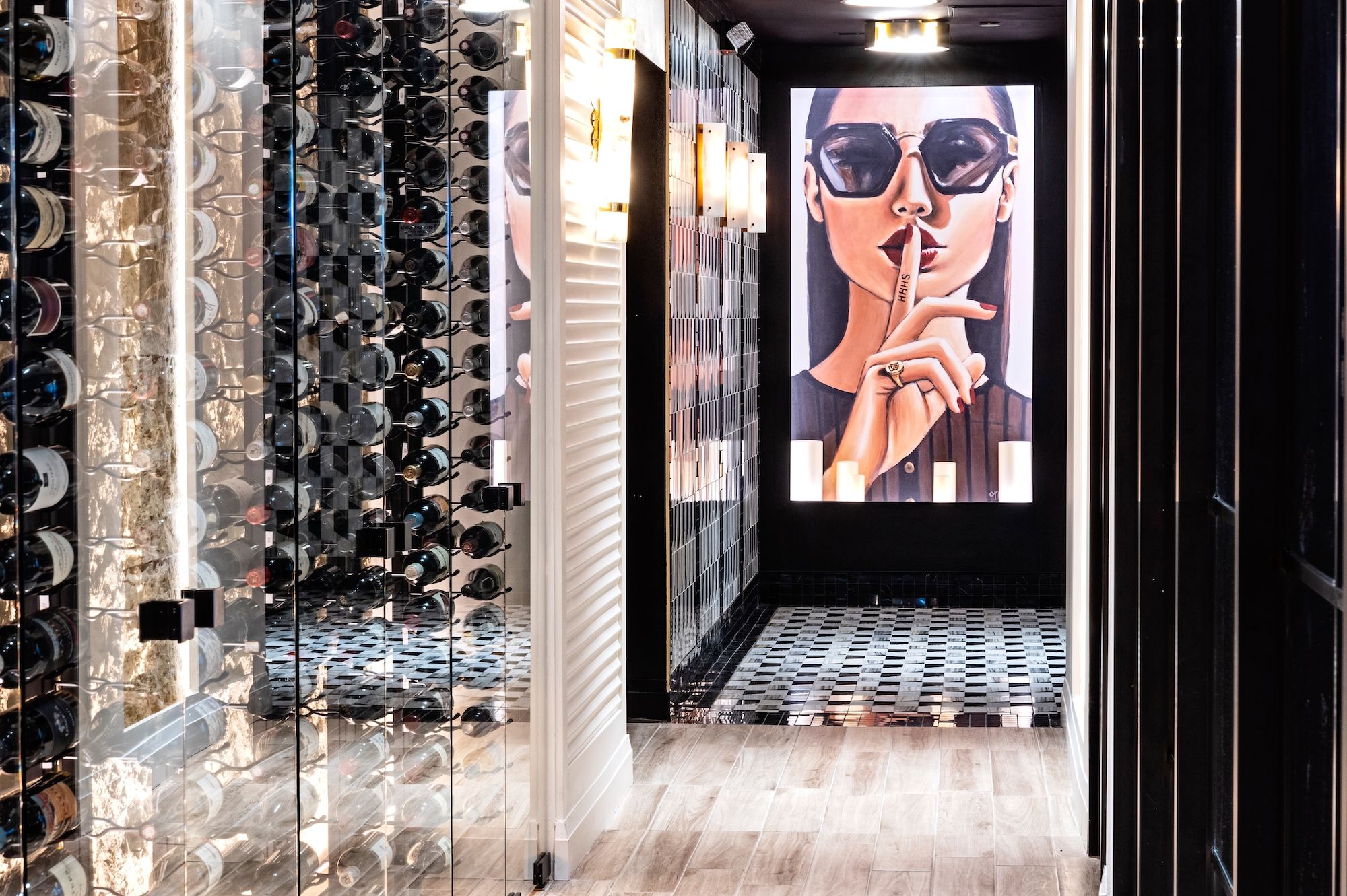 Hallway with glass wine racks on left, black and white checkered tile floor ahead, and illuminated artwork of woman with sunglasses and finger to lips saying "SHHH" on right wall.