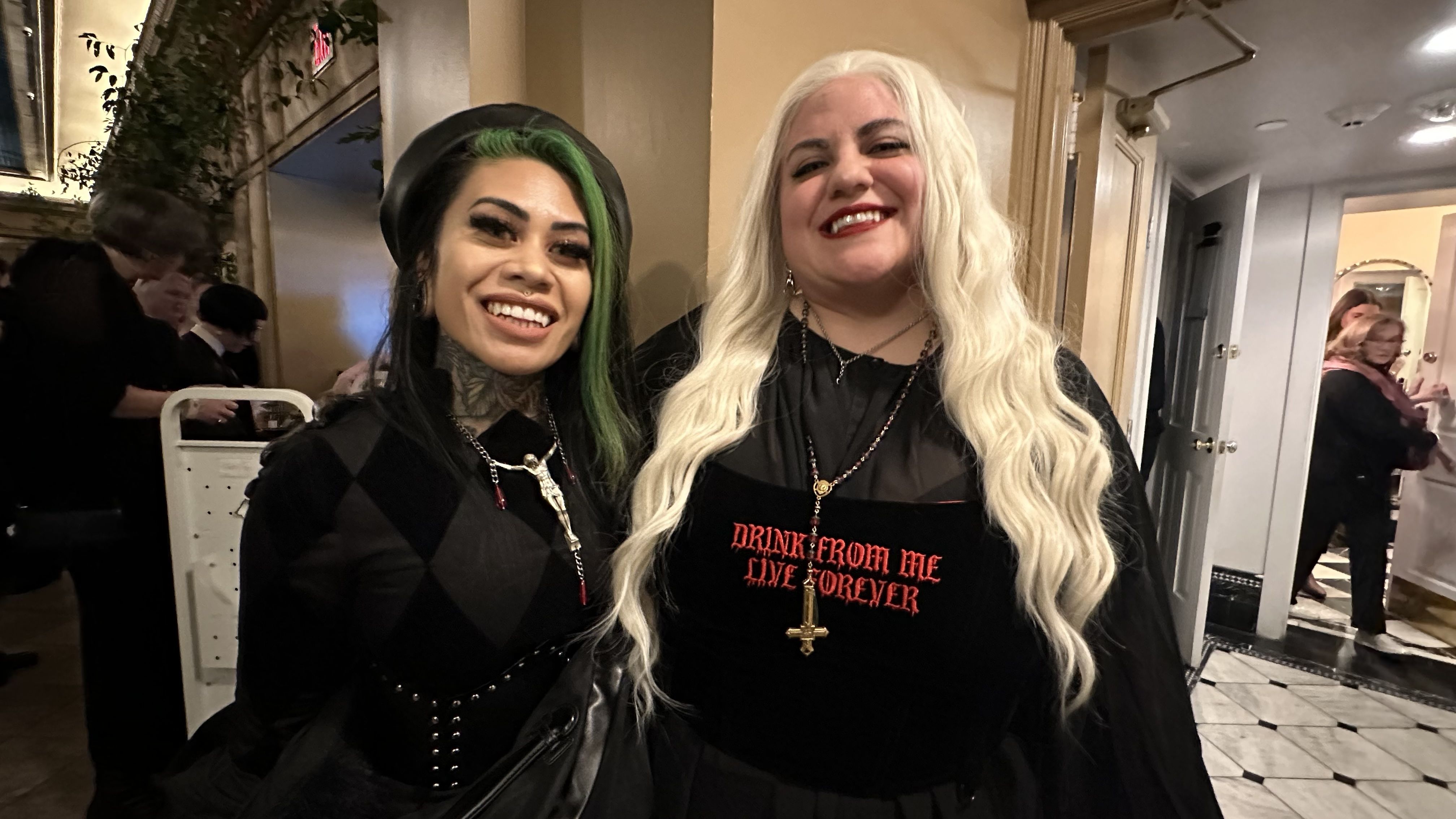Two women smiling indoors, one with long white hair and a shirt reading "DRINK FROM ME LIVE FOREVER" in red, the other with black and green hair, tattoos, and a black beret. Both wear black.