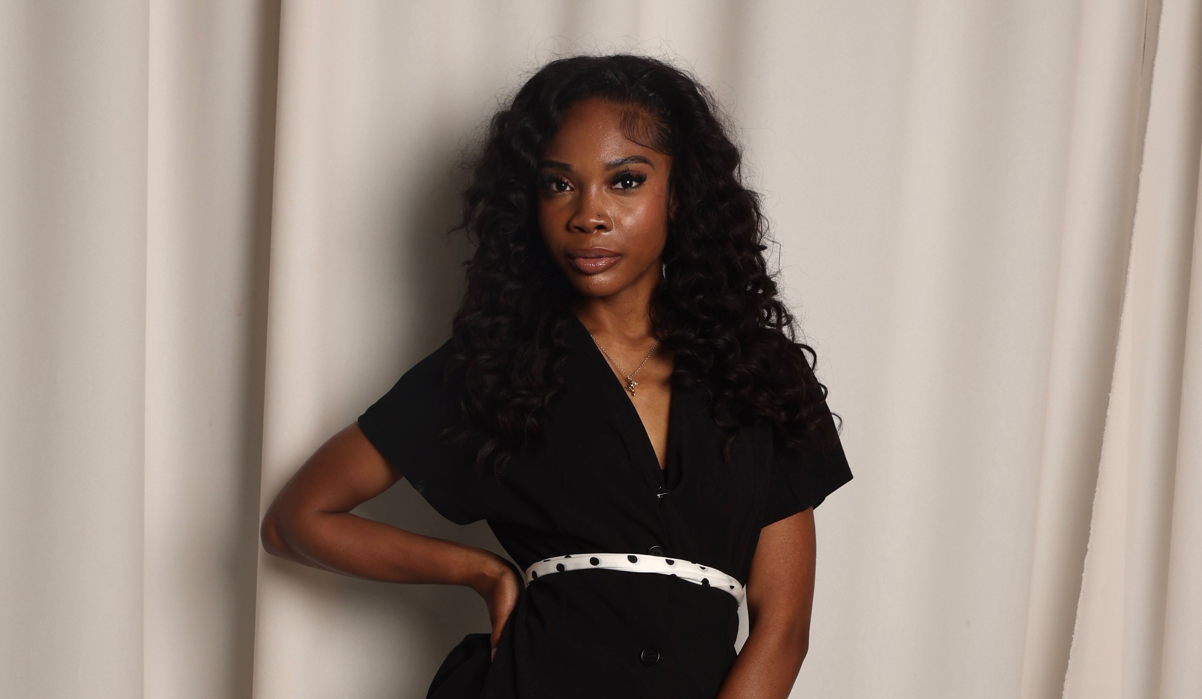 Portrait of a woman with long, curly black hair wearing a black dress and a white polka-dot belt, standing with one hand on her hip before beige curtains.