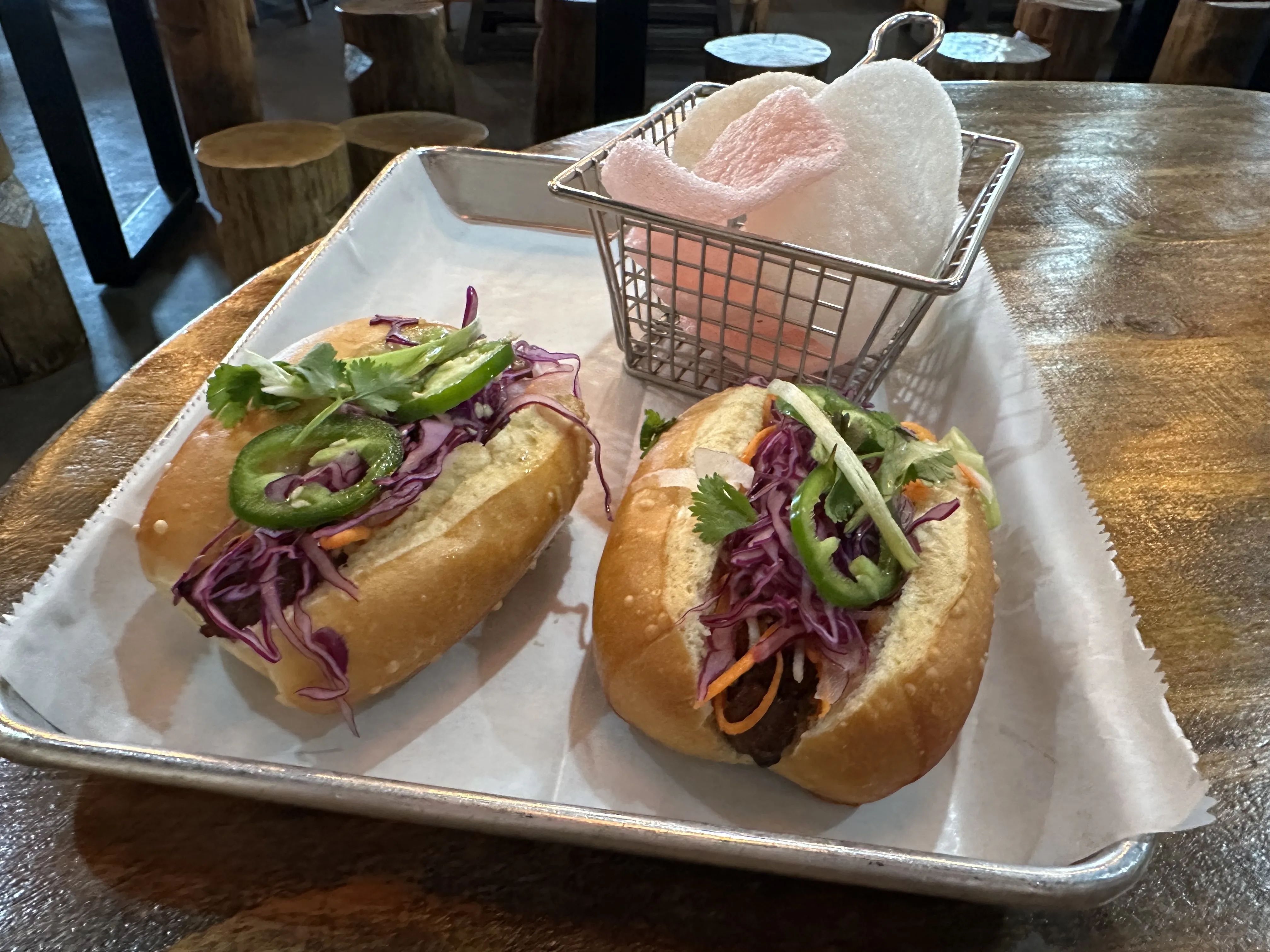 Two soft sandwich buns filled with purple cabbage slaw, jalapeño rounds, cilantro, and shredded carrots on a metal tray; a small wire basket holds white towels in the background.