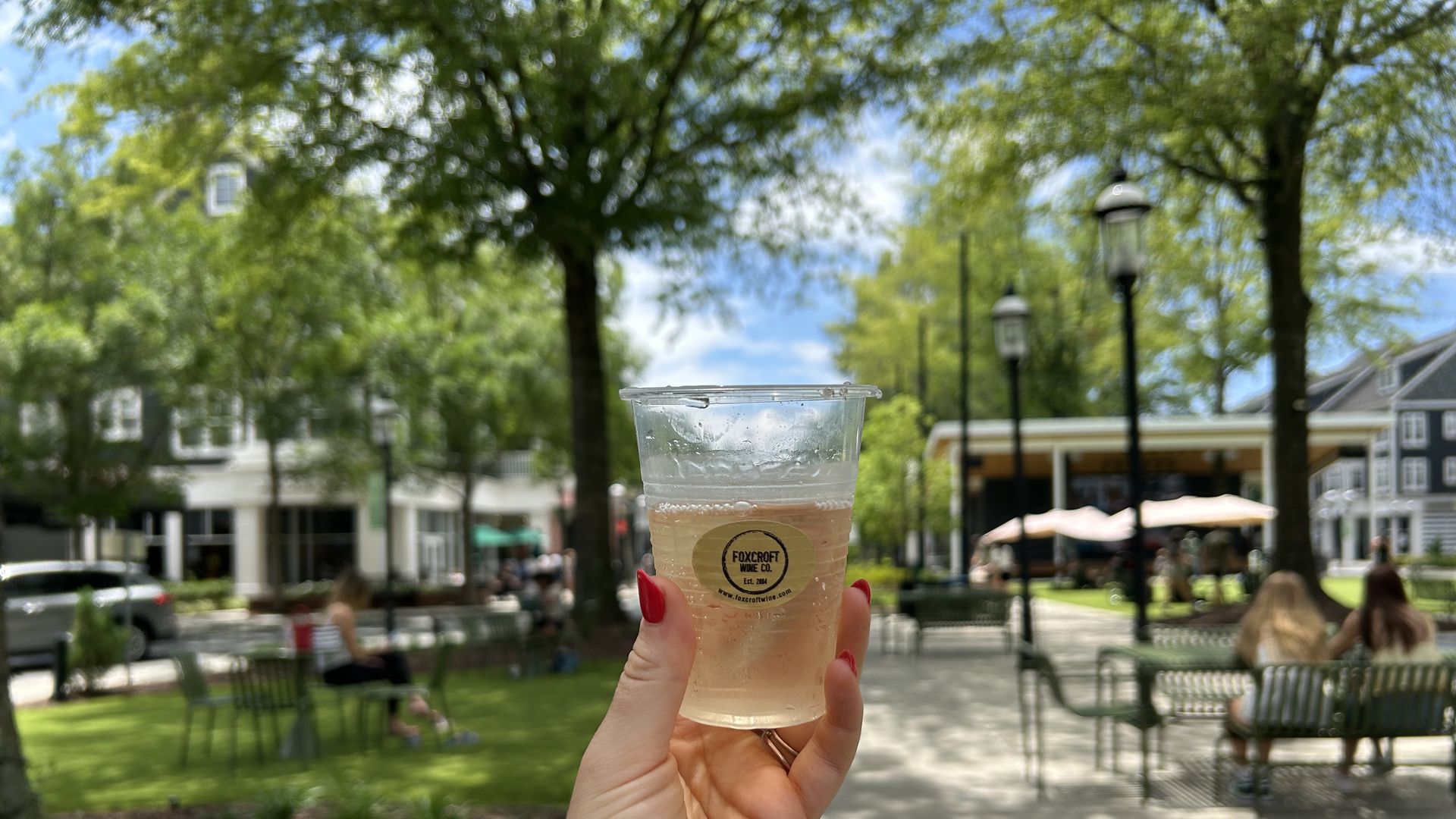 Hand with red nails holding a plastic cup of light-colored drink with Foxcroft Wine Co. label in an outdoor park area with benches, trees, and people on a sunny day.