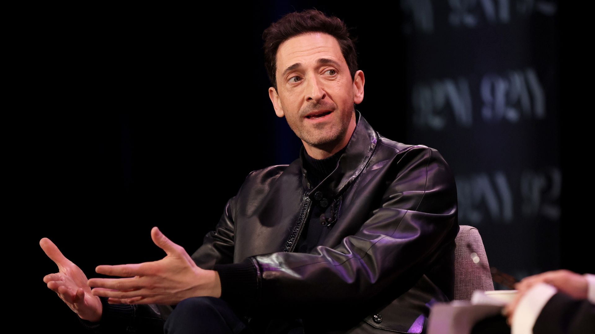 Adrien Brody in black jacket sits in interview and talks