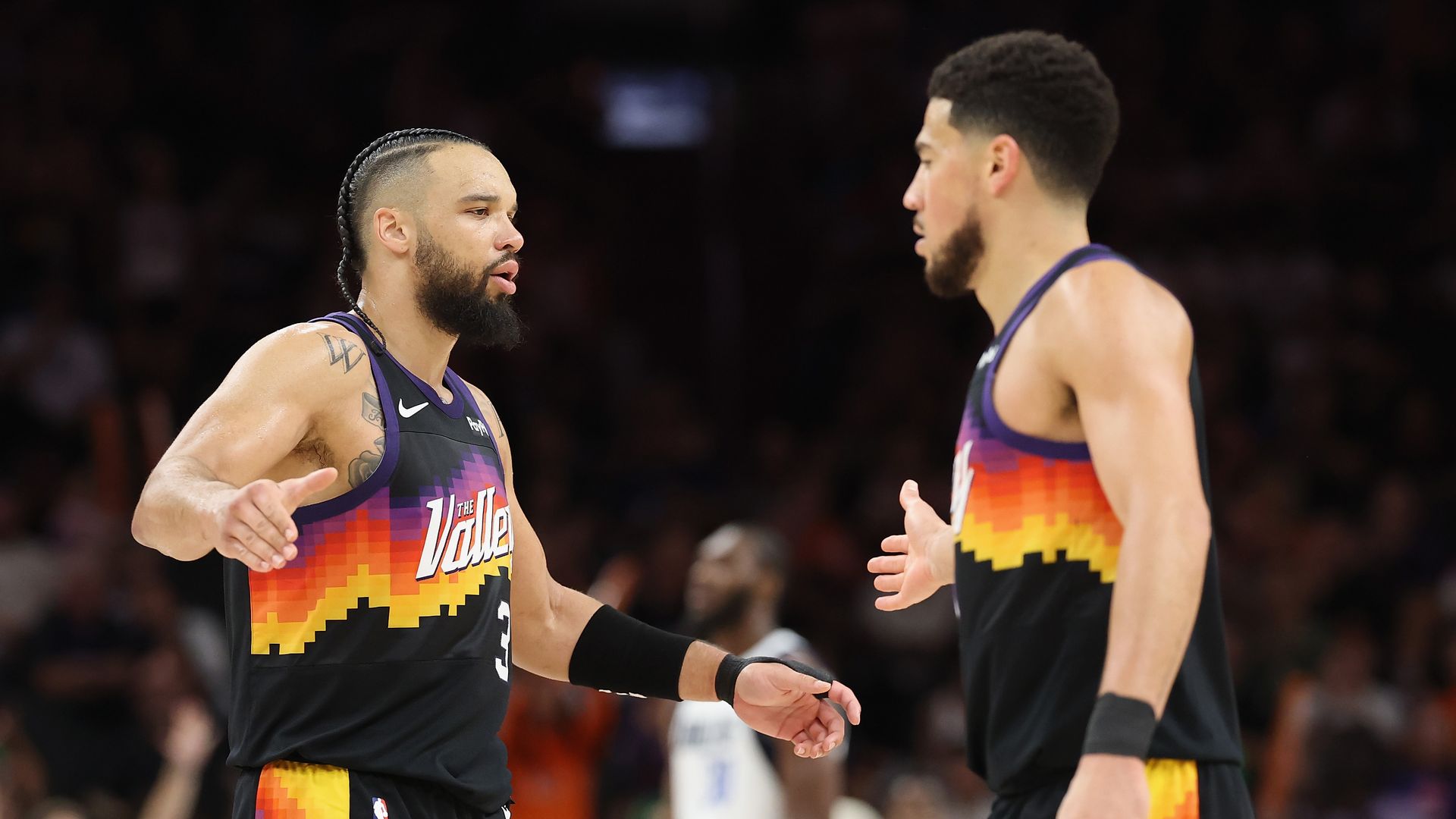 Two basketball players in black uniforms with orange, red and purple sunset designs wrapping around the chest and running down the sides of the legs slap hands on a basketball court, with fans seated in the background an opponent in a white uniform visible behind them.
