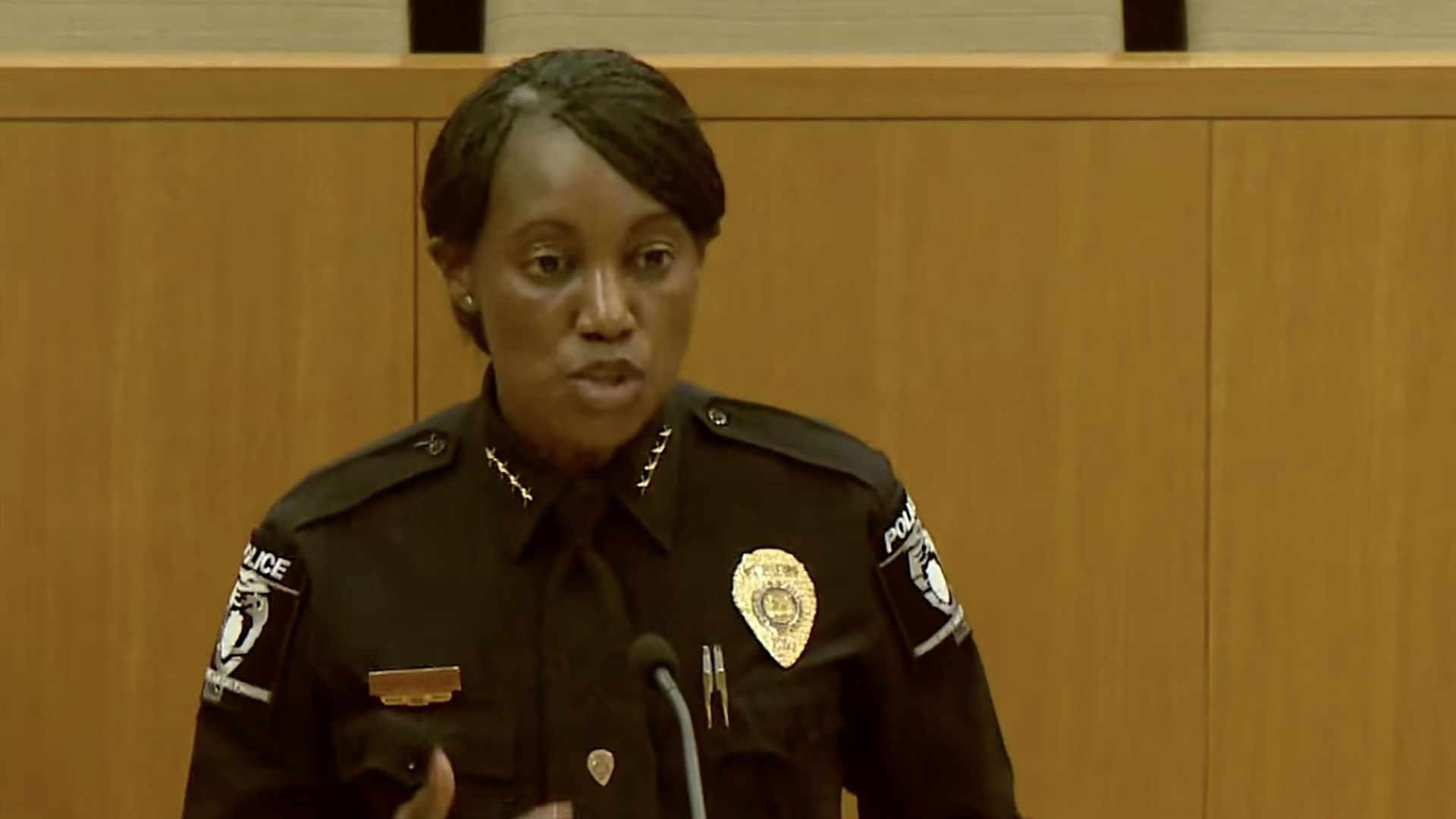 Female police officer in a dark uniform speaks into a microphone at a desk, with a gold badge on her chest, patches on her sleeves, and wood paneling behind her.