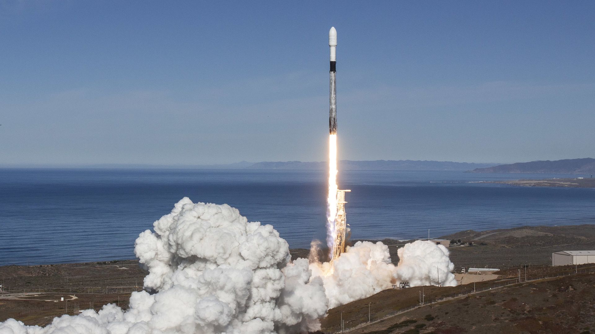 A SpaceX Falcon 9 rocket launching into a clear, blue sky with water and mountains on the horizon