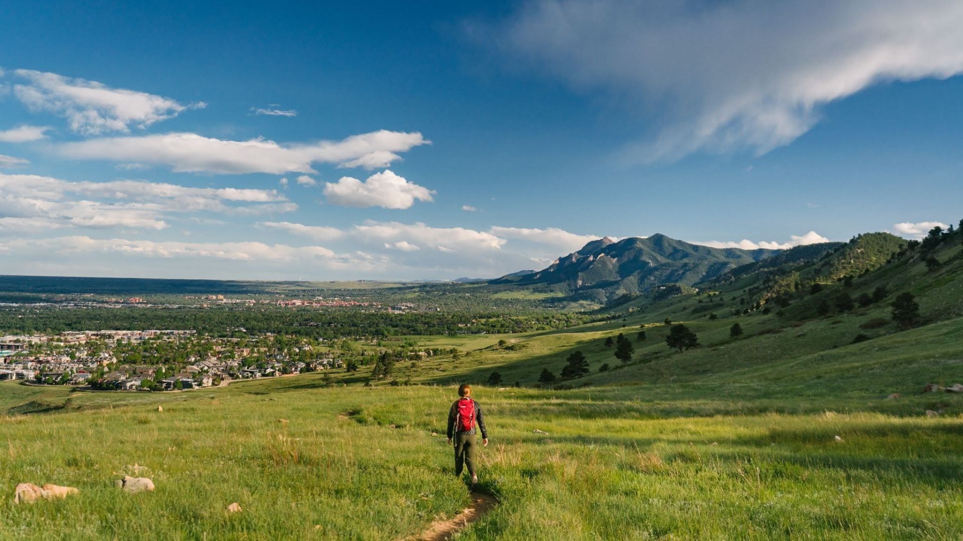 A hiker with a red backpack walks along a grassy hill toward distant mountains, with a valley town visible on the left under a bright blue sky with fluffy white clouds.