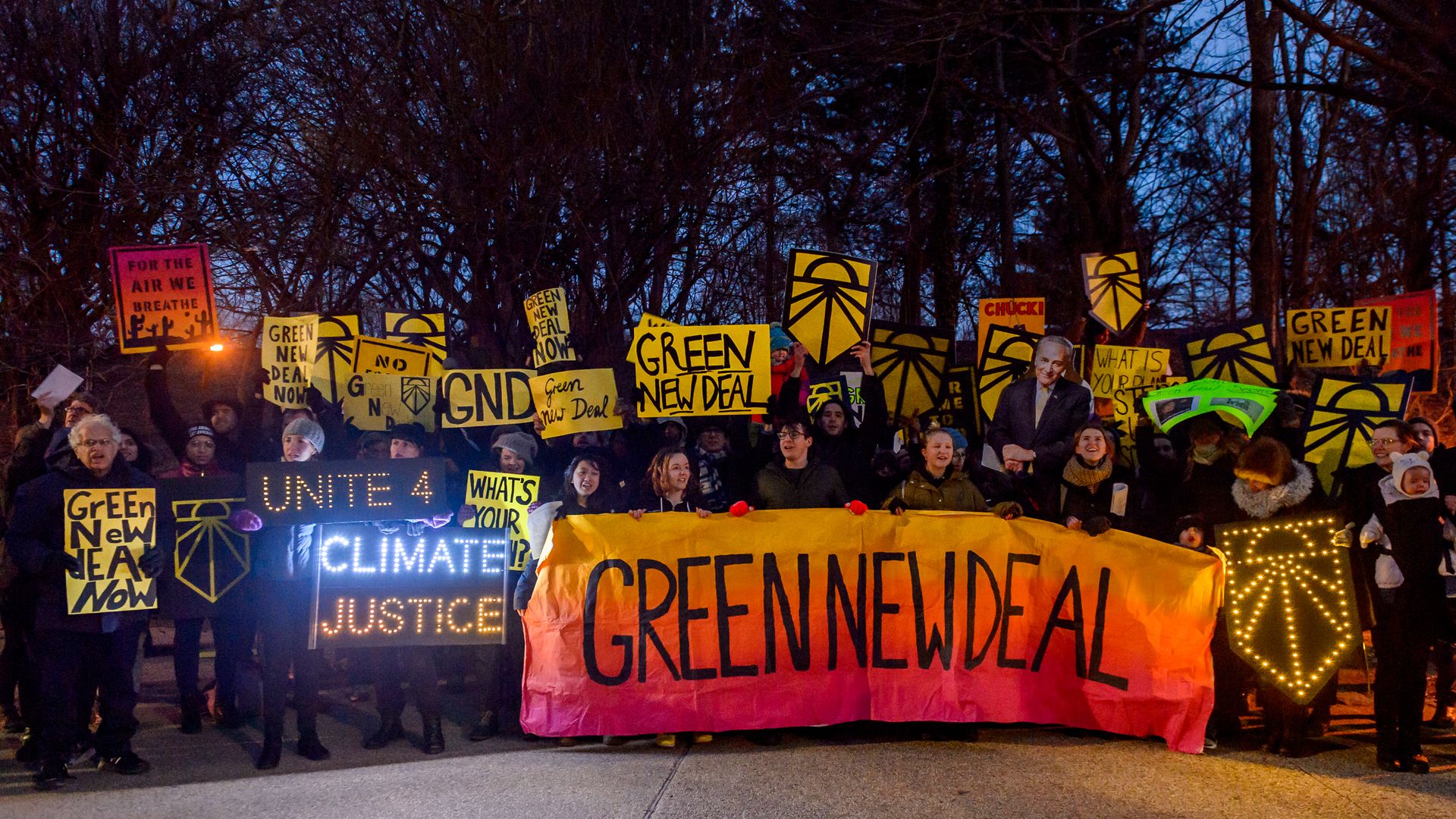 In this image, a line of demonstrators holding various signs support an orange banner that says "Green New Deal."