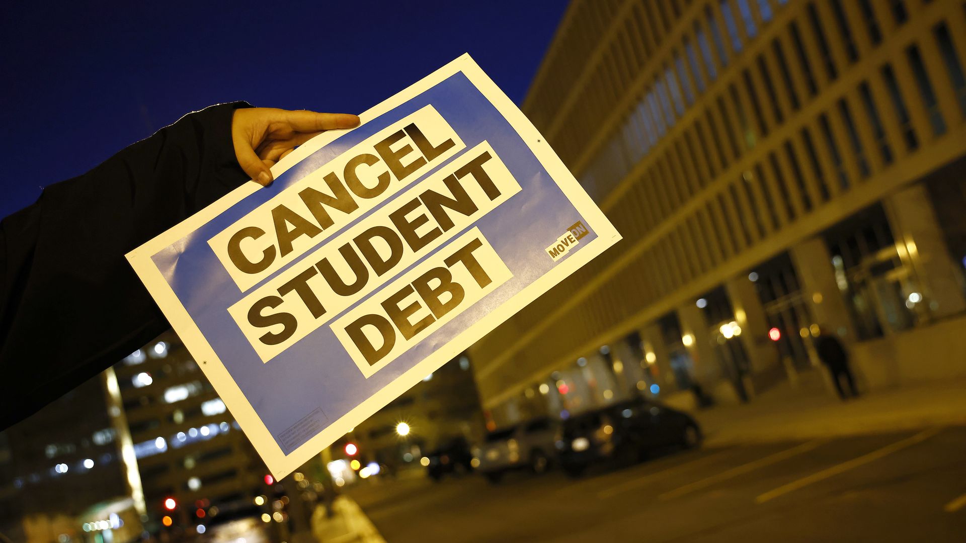 Photo of a hand holding a blue sign with black lettering that says "Cancel Student Debt"
