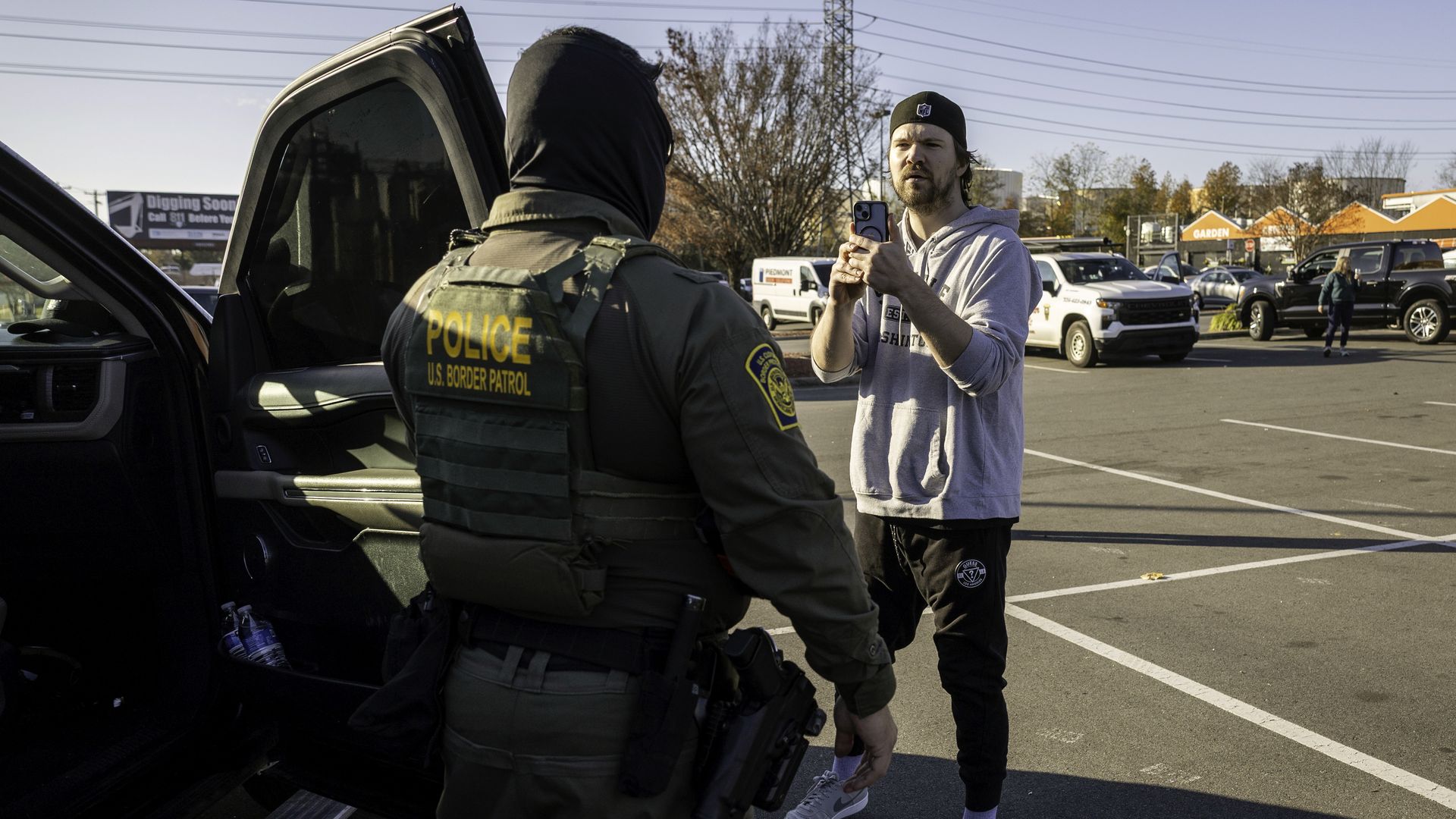 Man in gray hoodie and black cap films a U.S. Border Patrol officer wearing tactical gear and a balaclava near an open car door in a parking lot on a sunny day.