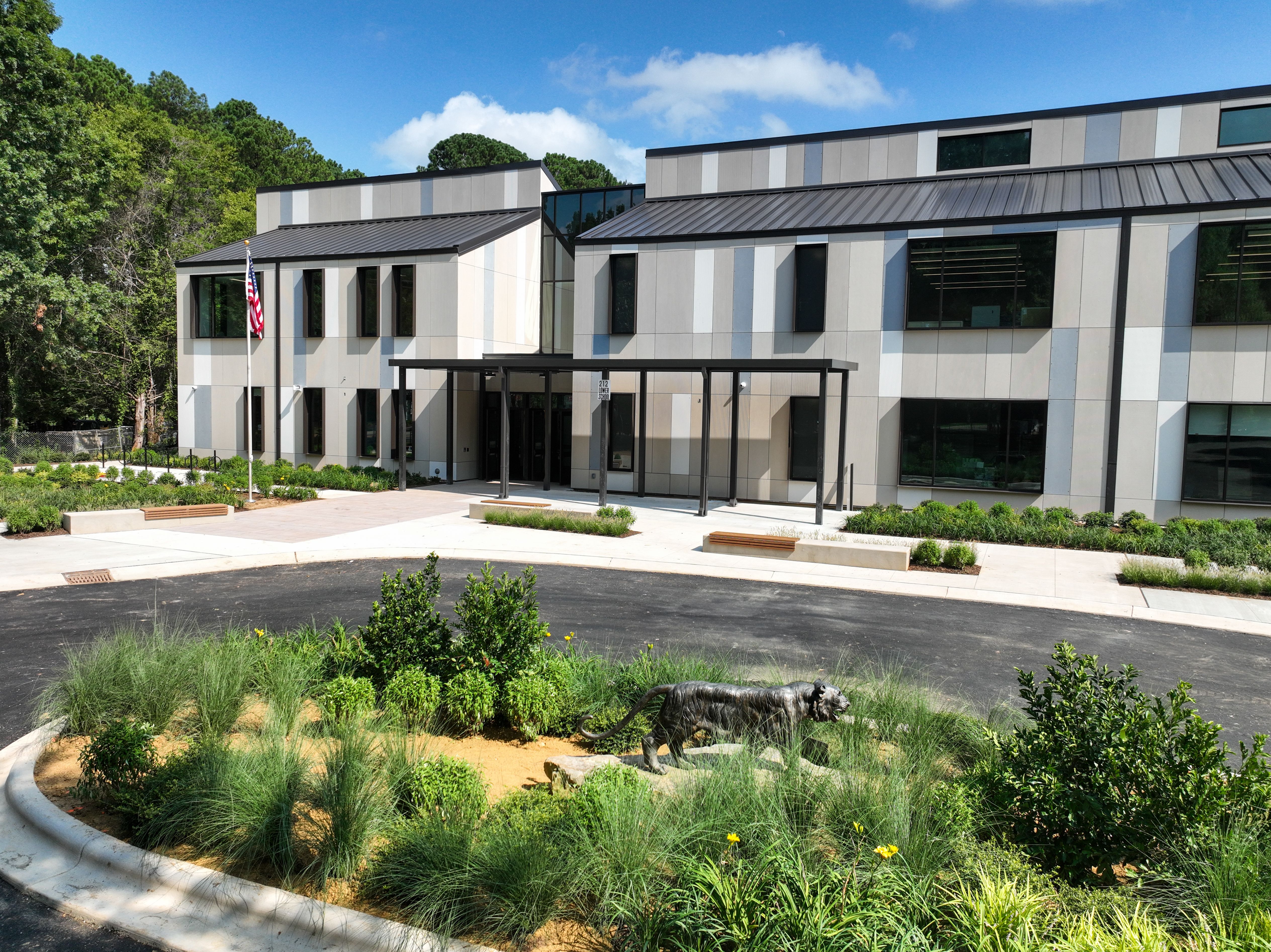 Charlotte Preparatory School's modern two-story building with gray panel exterior, a covered entrance, an American flag, and a roundabout featuring a tiger statue surrounded by green plants under a blue sky.