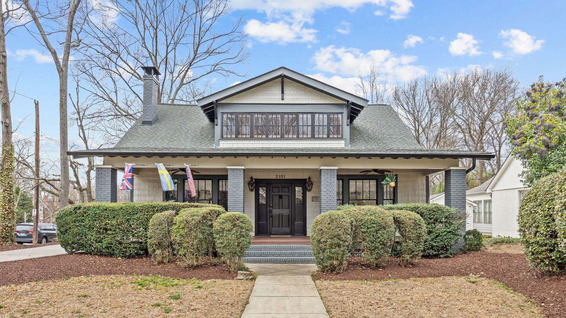 Gray roofed house with black door and columns, bushes in front, three flags on porch, leafless trees behind, and partly cloudy blue sky.