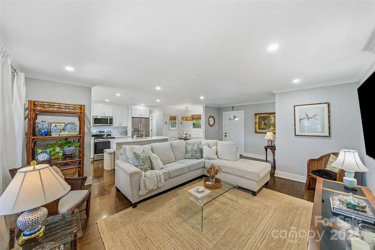 Bright open living room with large light gray sectional, glass coffee table, beige woven rug, wooden floor, kitchen in background, and decorative shelves and lamps.
