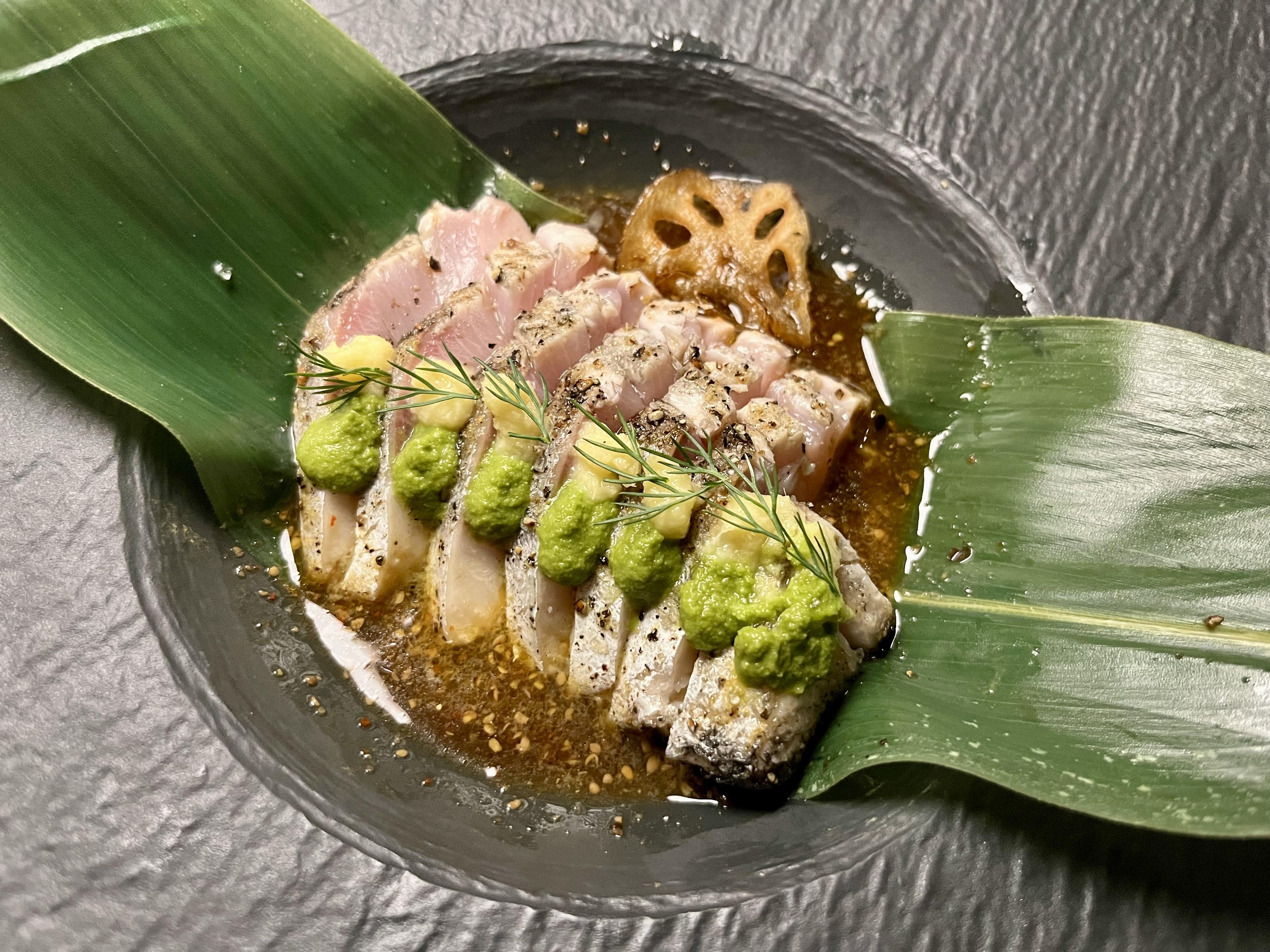 Fish sits inside a bowl of oil and inside a large green leaf, and is topped with dill and ginger.