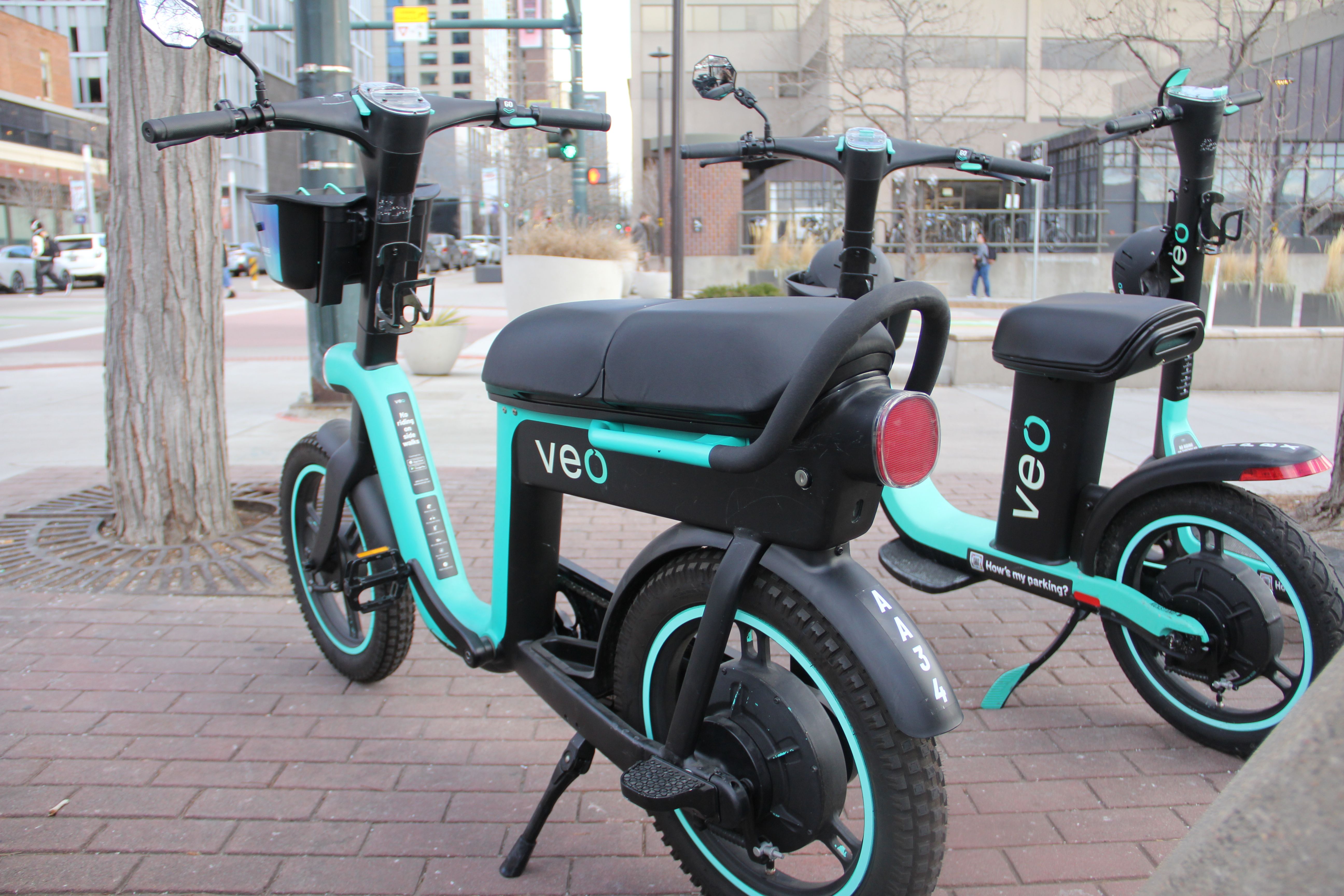 Two teal and black shared electric scooters labeled "veo" parked on a city sidewalk near a tree and urban buildings.