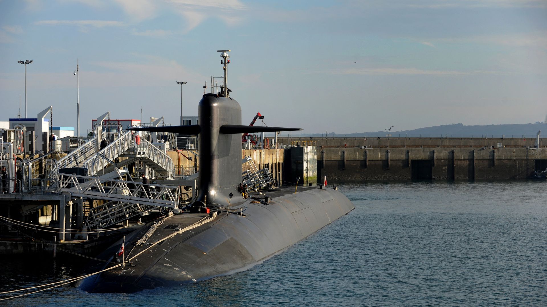 Black military submarine docked at a harbor with access gangways and a calm sea under a partly cloudy sky.