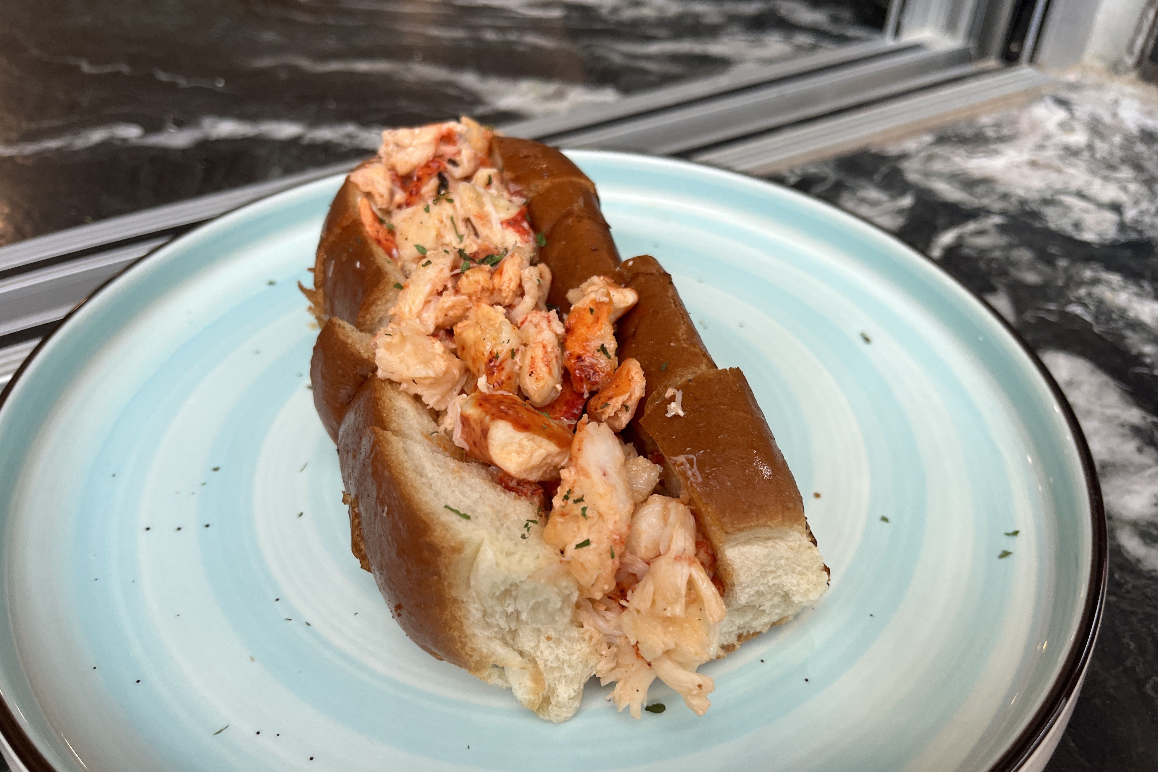 Lobster roll with chunky lobster meat in a light mayo dressing, garnished with parsley, inside a toasted brown bun on a pale blue plate; dark granite counter in the background.