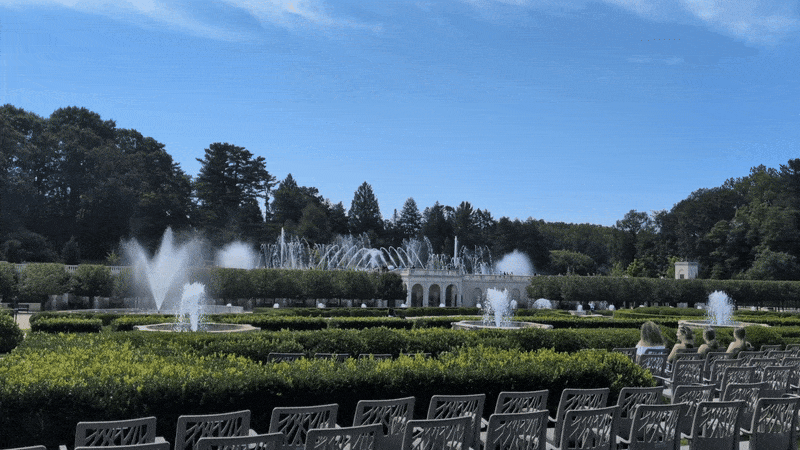 Outdoor garden with multiple water fountains spraying in a circular pattern, surrounded by neatly trimmed green hedges, white chairs, and a few people sitting under a clear blue sky.