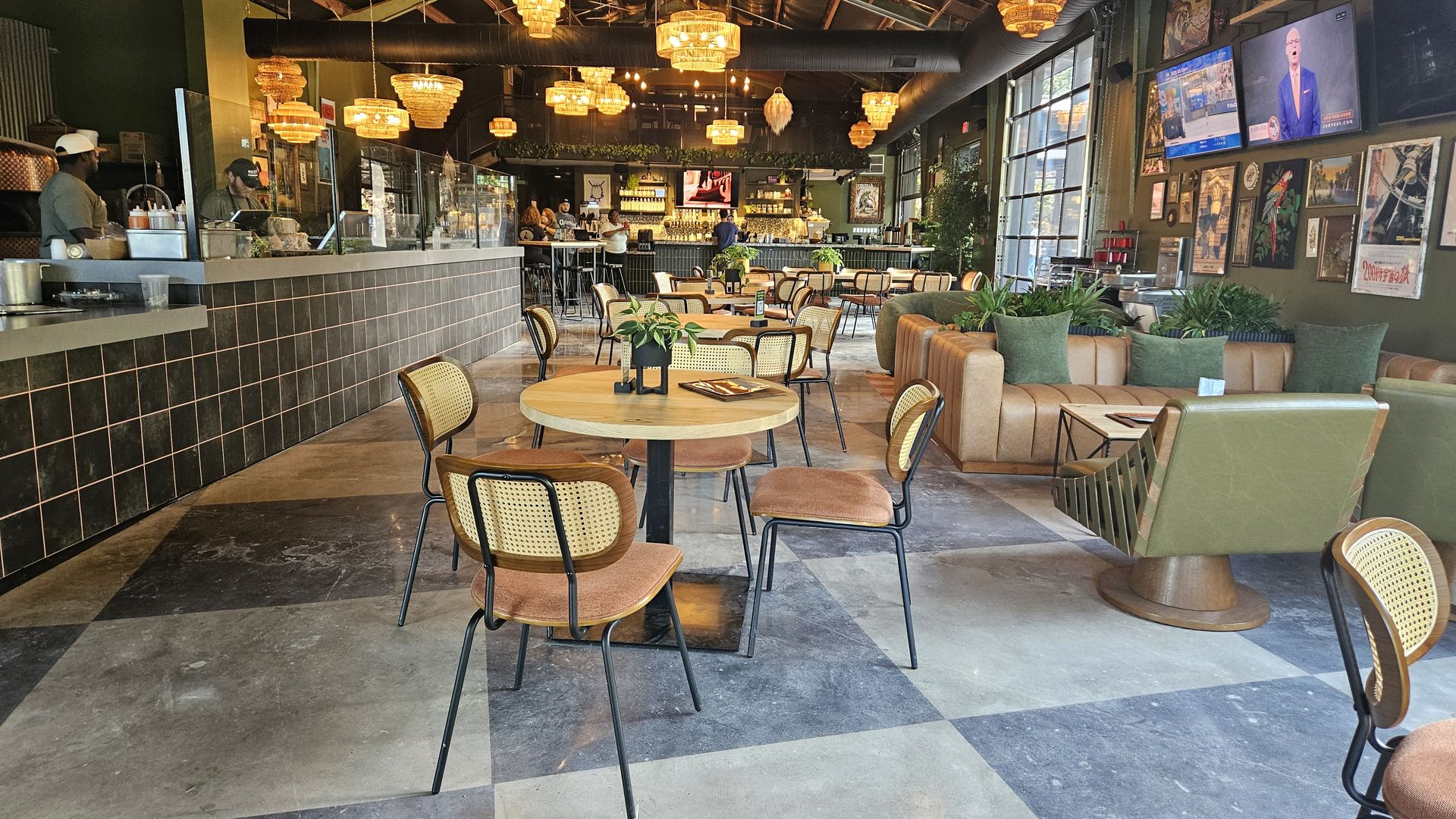 Interior of a modern brewery with wooden tables, cane-back chairs, pendant lights, and a bar in the background. Green leather chairs and a brown sofa with green pillows are on the right.