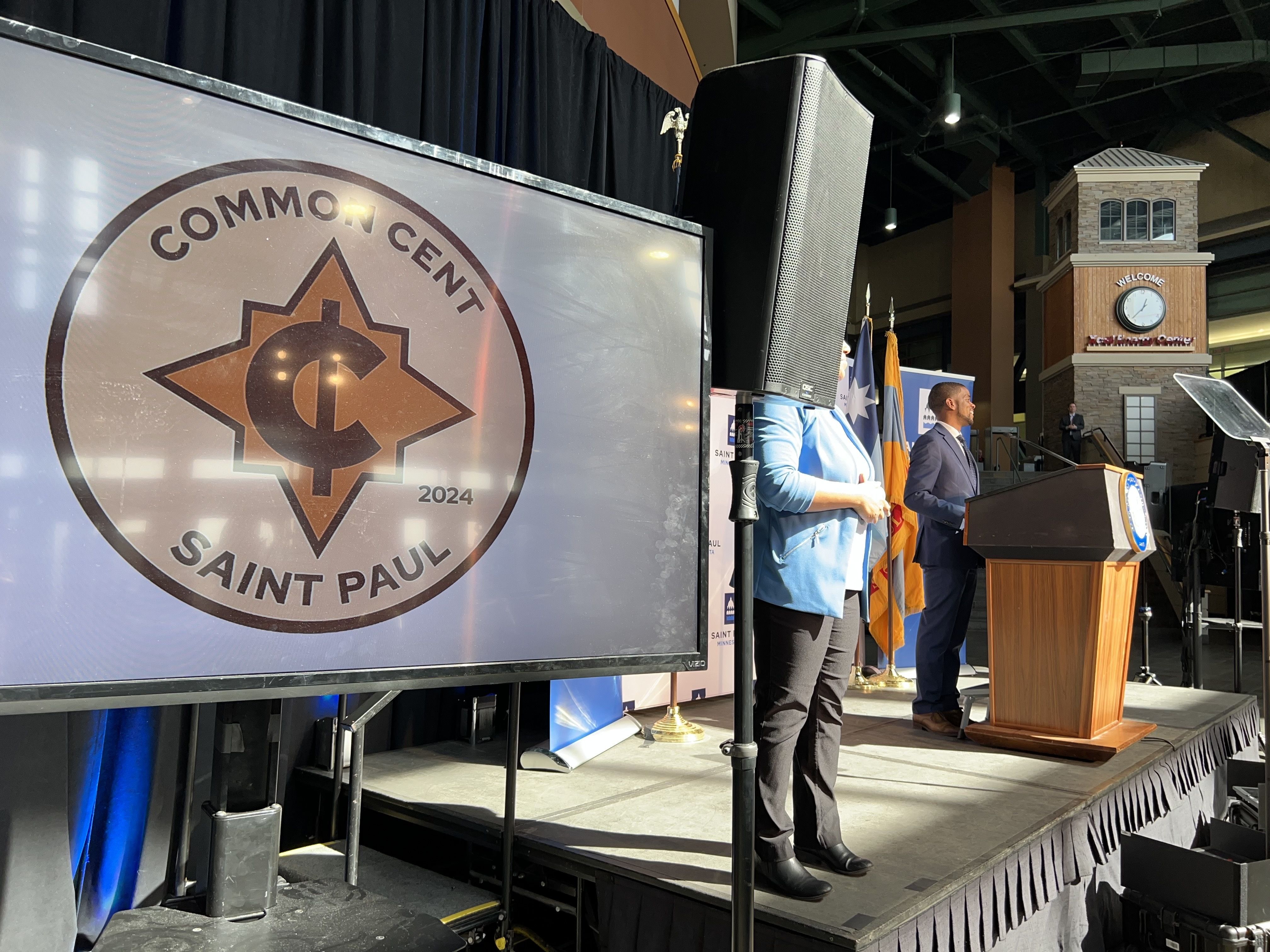 A screen with a circular logo reading "Common Cent: Saint Paul, 2024" displayed next to a politician's podium where a Black man in a business suit stands in front of a teleprompter.