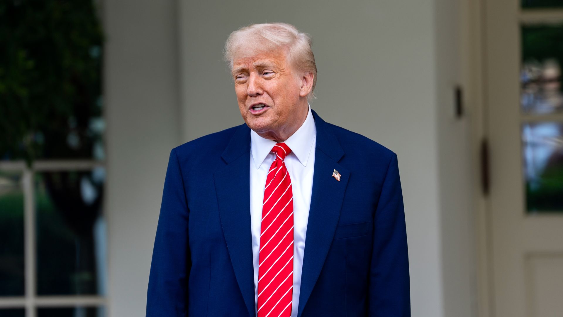 Donald Trump wearing a blue suit and red tie, standing in front of a white wall and doors.