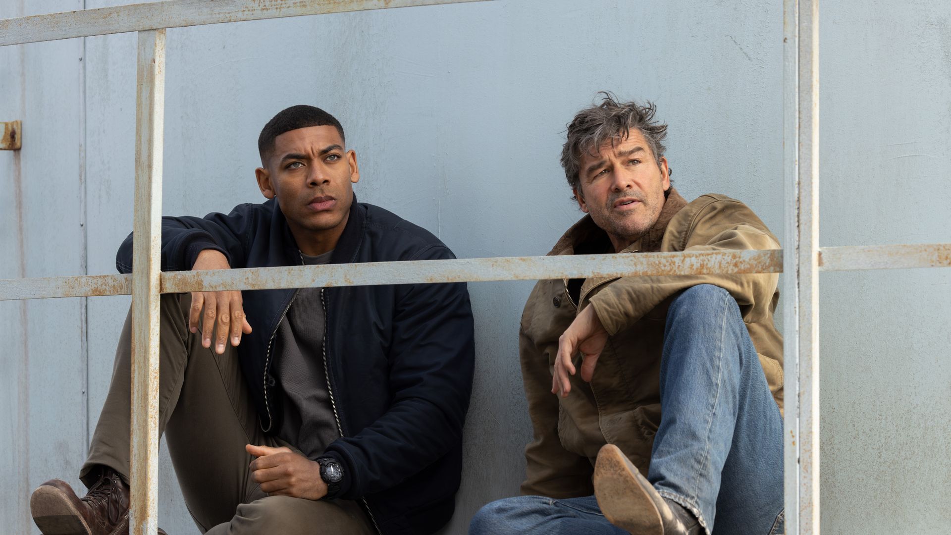 Aaron Pierre and Kyle Chandler sit on a rusted metal railing against a pale blue wall, looking off to the side.