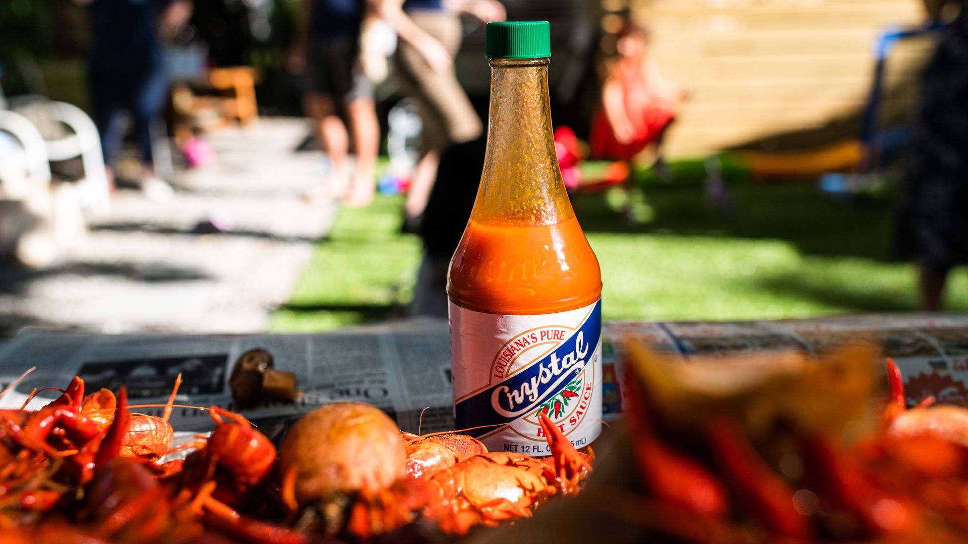 A bottle of Crystal hot sauce with a green cap is surrounded by boiled crawfish on a newspaper-covered table outdoors, with blurred people and grass in the background.