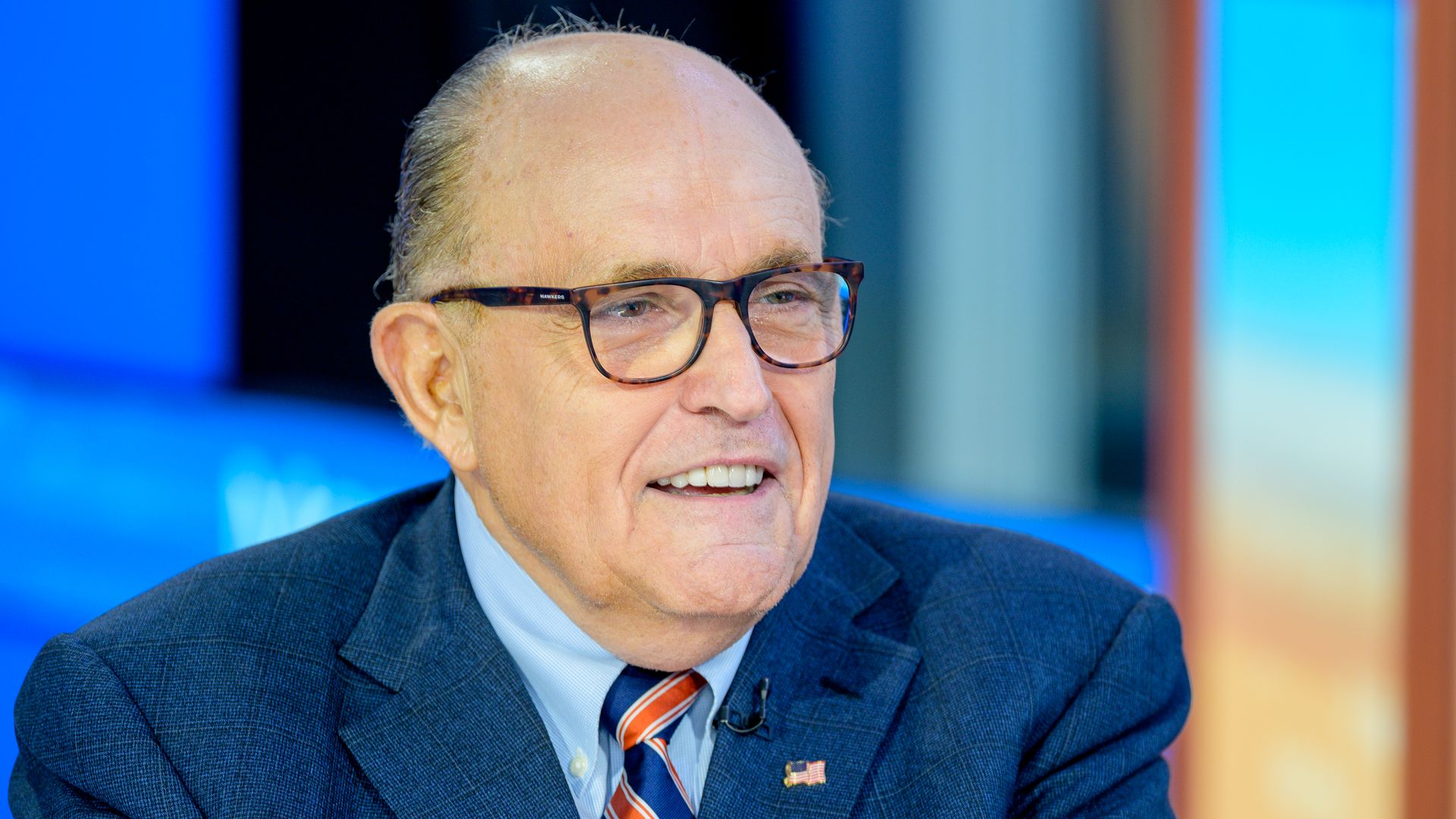 In this image, Giuliani wears a suit and glasses and smiles