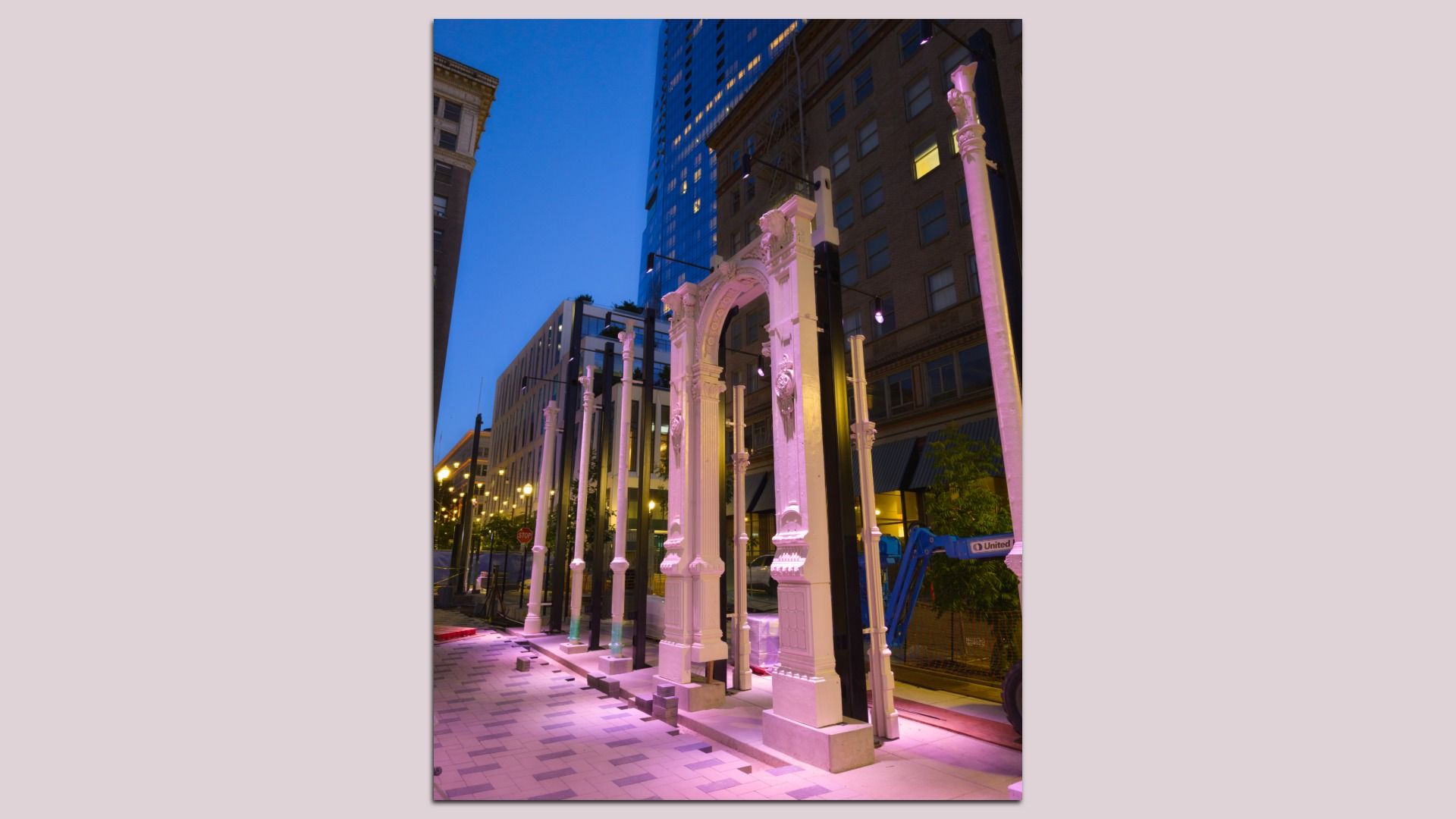 Row of ornate white arches and columns lit pink along a city street at dusk; modern glass and brick buildings rise behind, with a patterned sidewalk in the foreground.