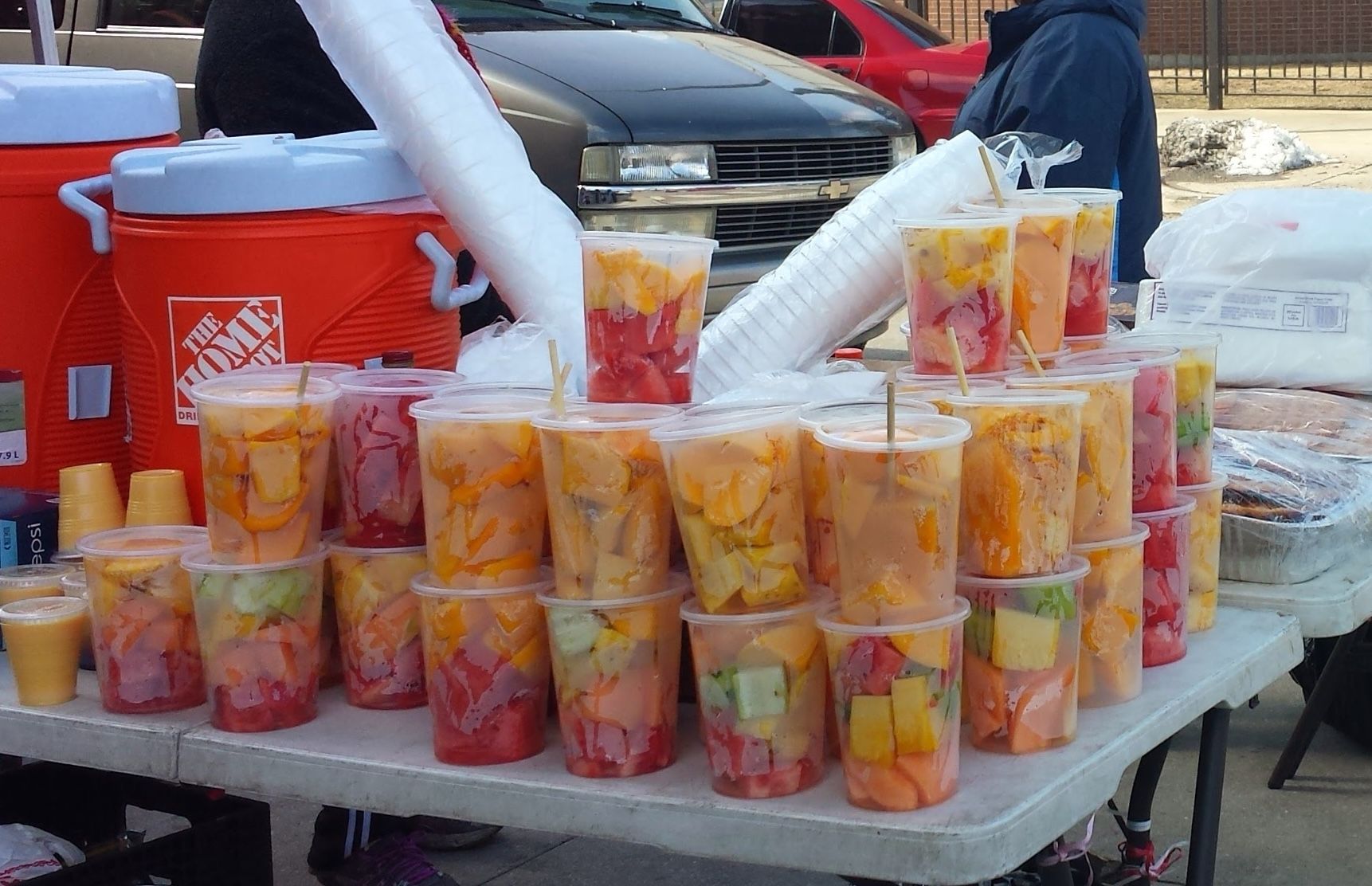 Row of clear cups filled with colorful fruit salad on a table, with a red Home Depot cooler and parked cars in the background.