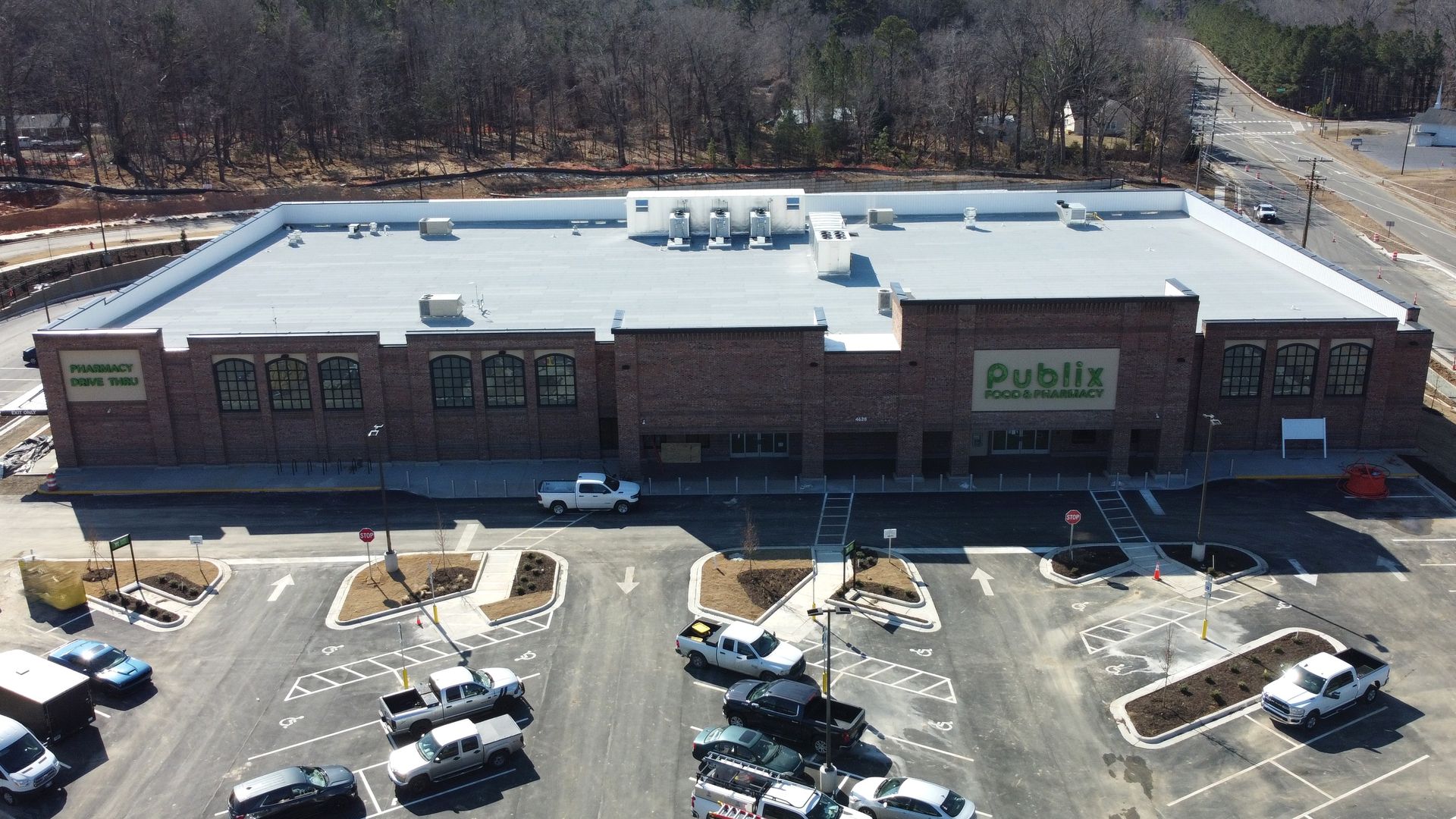 Aerial view of a Publix Food & Pharmacy with a large white roof and brick facade, surrounded by a mostly empty parking lot with several trucks and cars, trees and roads nearby.