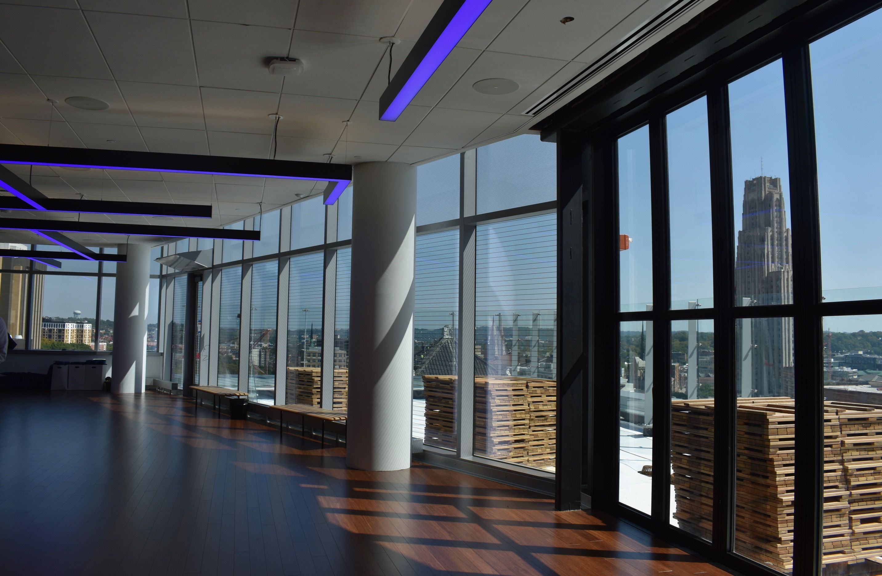 Modern indoor space with dark wood floors, white columns, large windows, and blue LED ceiling lights; cityscape with tall buildings visible through the windows on a clear day.