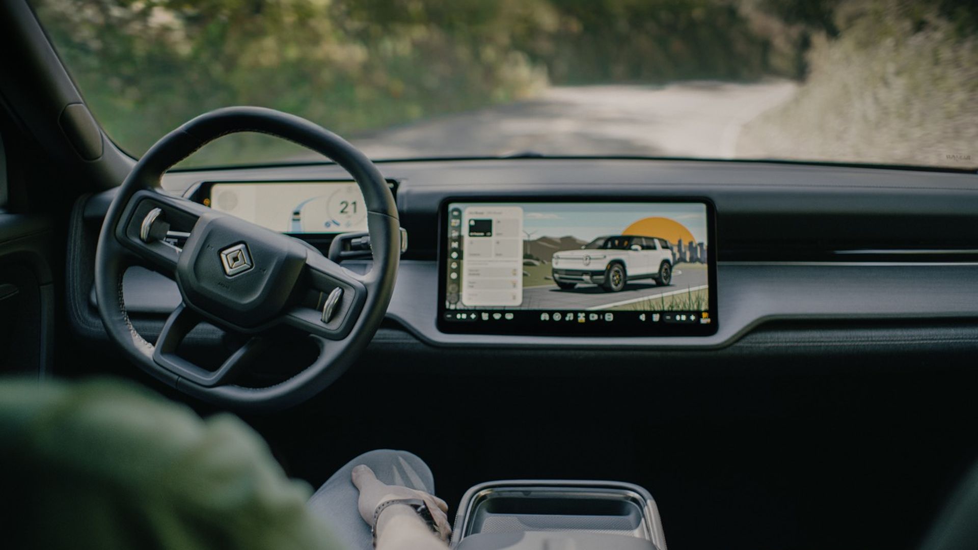 Car interior with a left-hand steering wheel and a wide touchscreen showing a white SUV near an orange sunset; passenger leg in foreground, road and greenery visible outside.