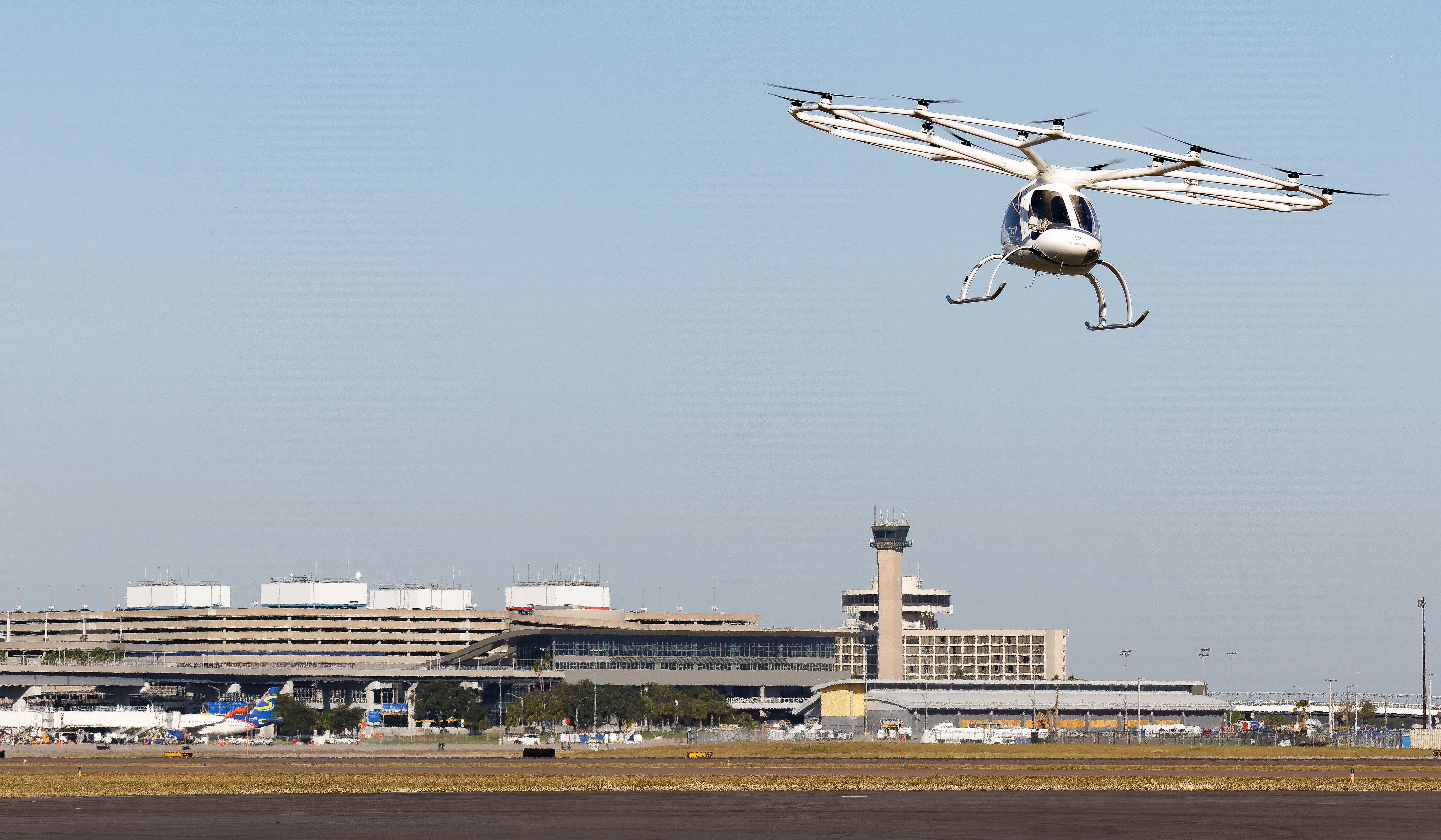 White gyroplane with a large circular rotor ring and skid landing gear flies over an airport, with a control tower and terminal building in the background on a clear day.