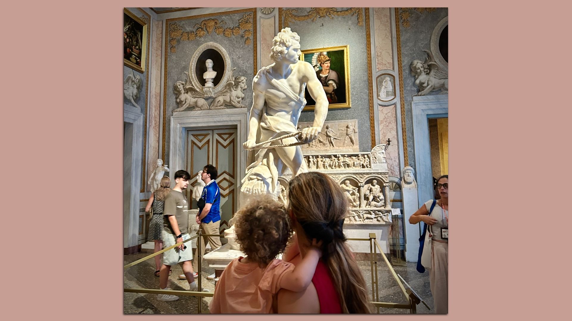 White marble statue of a muscular man gripping a rope in a lavish museum hall with gold-trimmed walls, ornate reliefs and a woman and a child facing the statue.