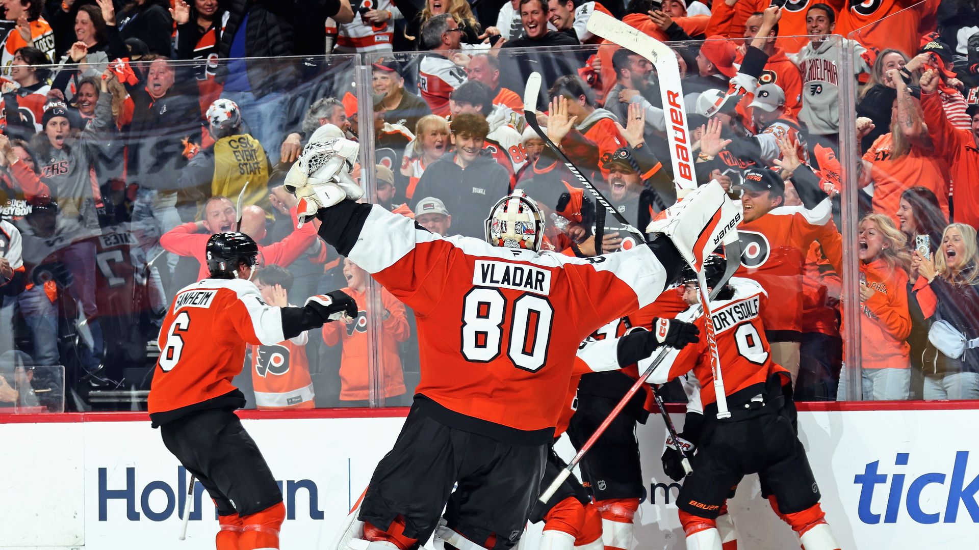 Hockey players in bright orange celebrate on the rink; goalie Vladar (#80) raises arms as a packed, cheering crowd in orange jerseys cheers behind the glass.