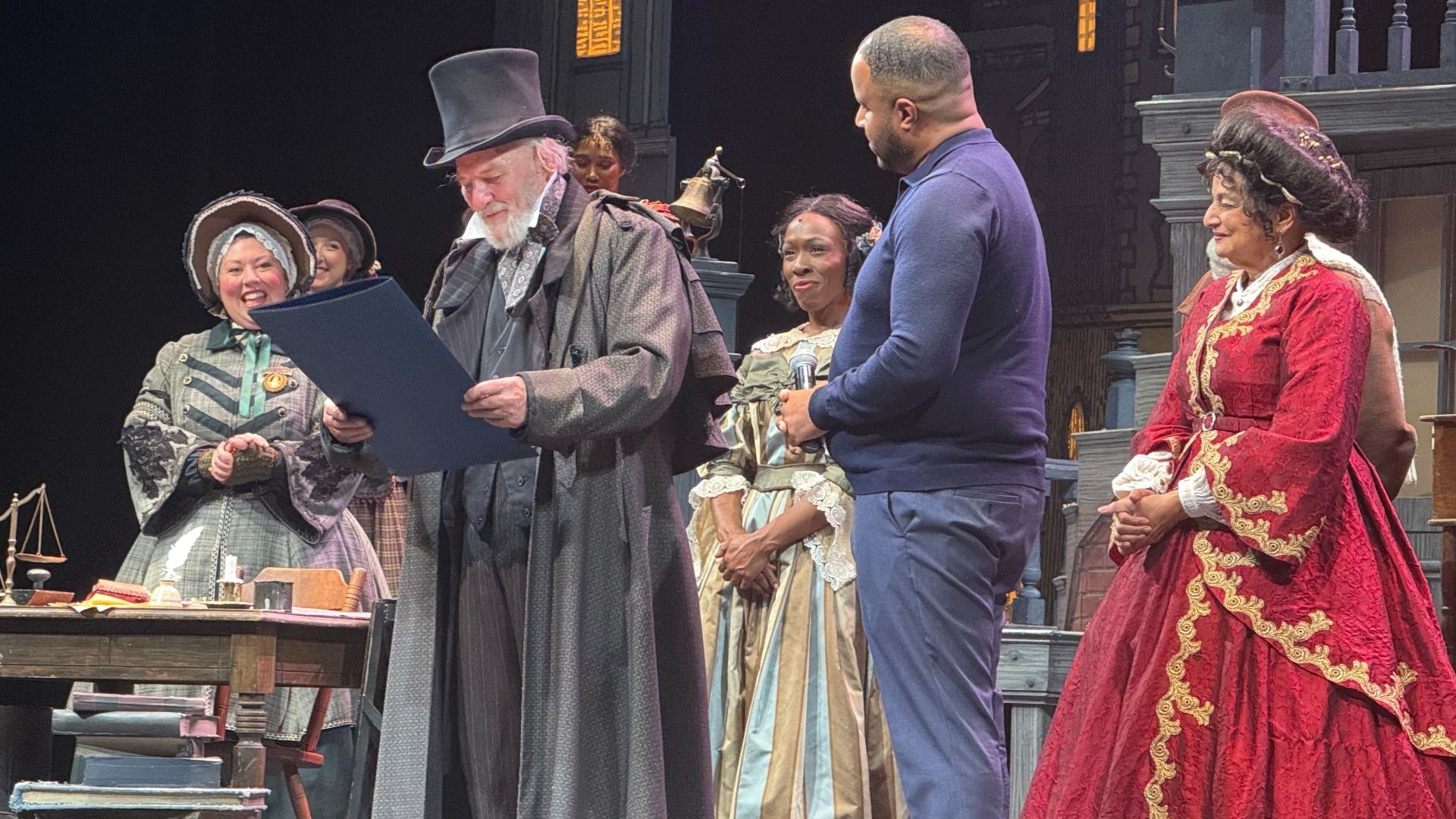 Actors in Victorian-era costumes on stage, one man in a top hat reading a document, surrounded by others in period dresses and a modern man in a blue sweater holding a microphone.