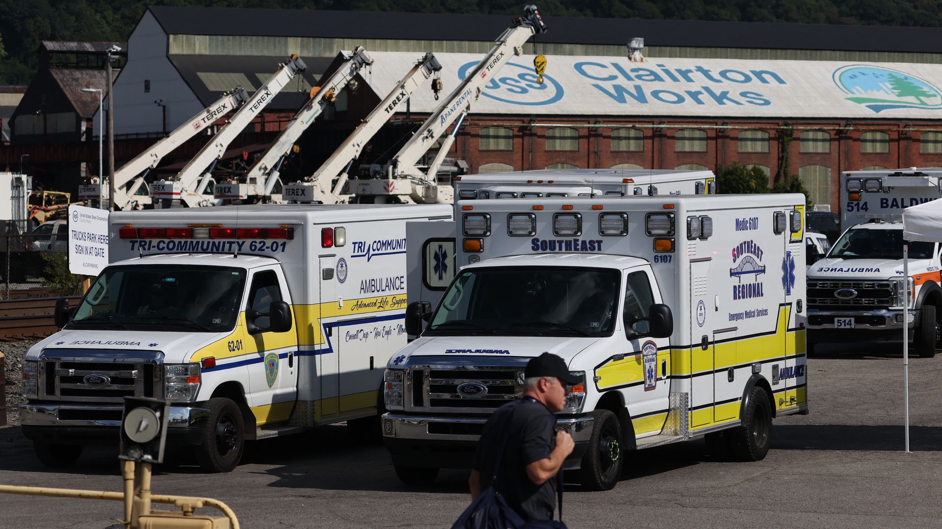 Parked white and yellow ambulances with a man carrying a bag walking in front, industrial building and cranes in the background.