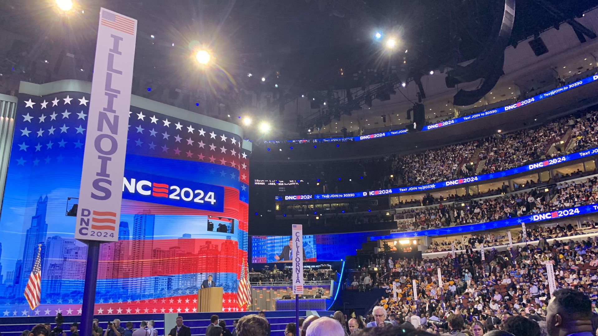 Crowd at DNC 2024 event in arena with stage, large screens showing red, white, blue patriotic design, Illinois and California delegate signs visible, people taking photos and watching