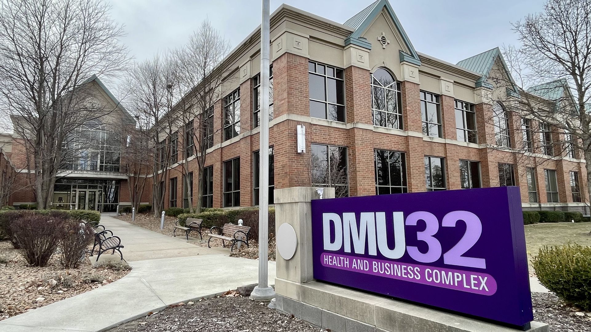 Red brick building with large windows and green roofs behind a purple sign reading "DMU32 Health and Business Complex" on a cloudy day with leafless trees and benches nearby.