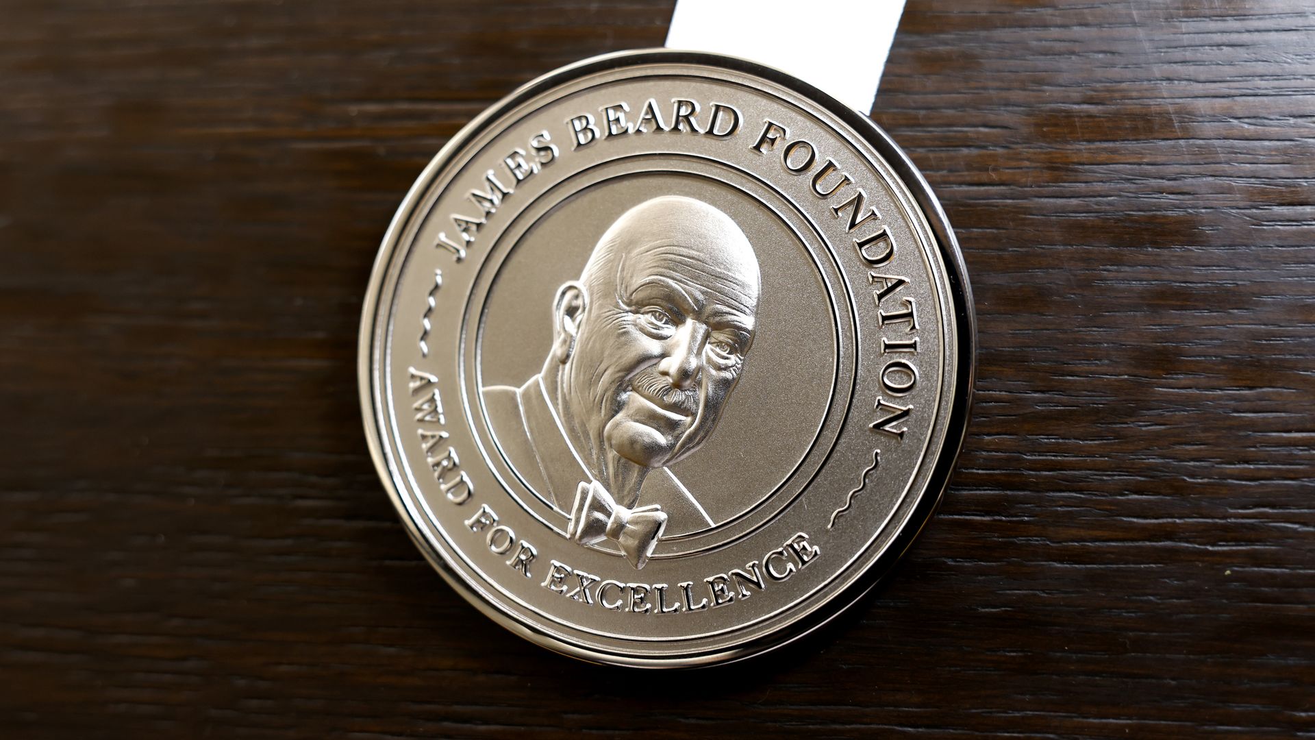 Close-up of a silver James Beard Foundation medal with a portrait and raised lettering, attached to a white ribbon, on a dark wooden surface.