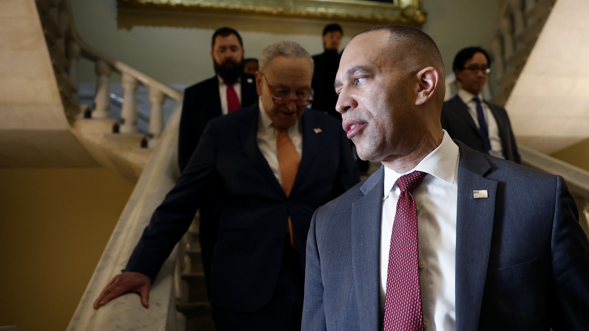 Hakeem Jeffries, wearing a gray suit and standing at the bottom of a staircase which Chuck Schumer and aides and security are descending.