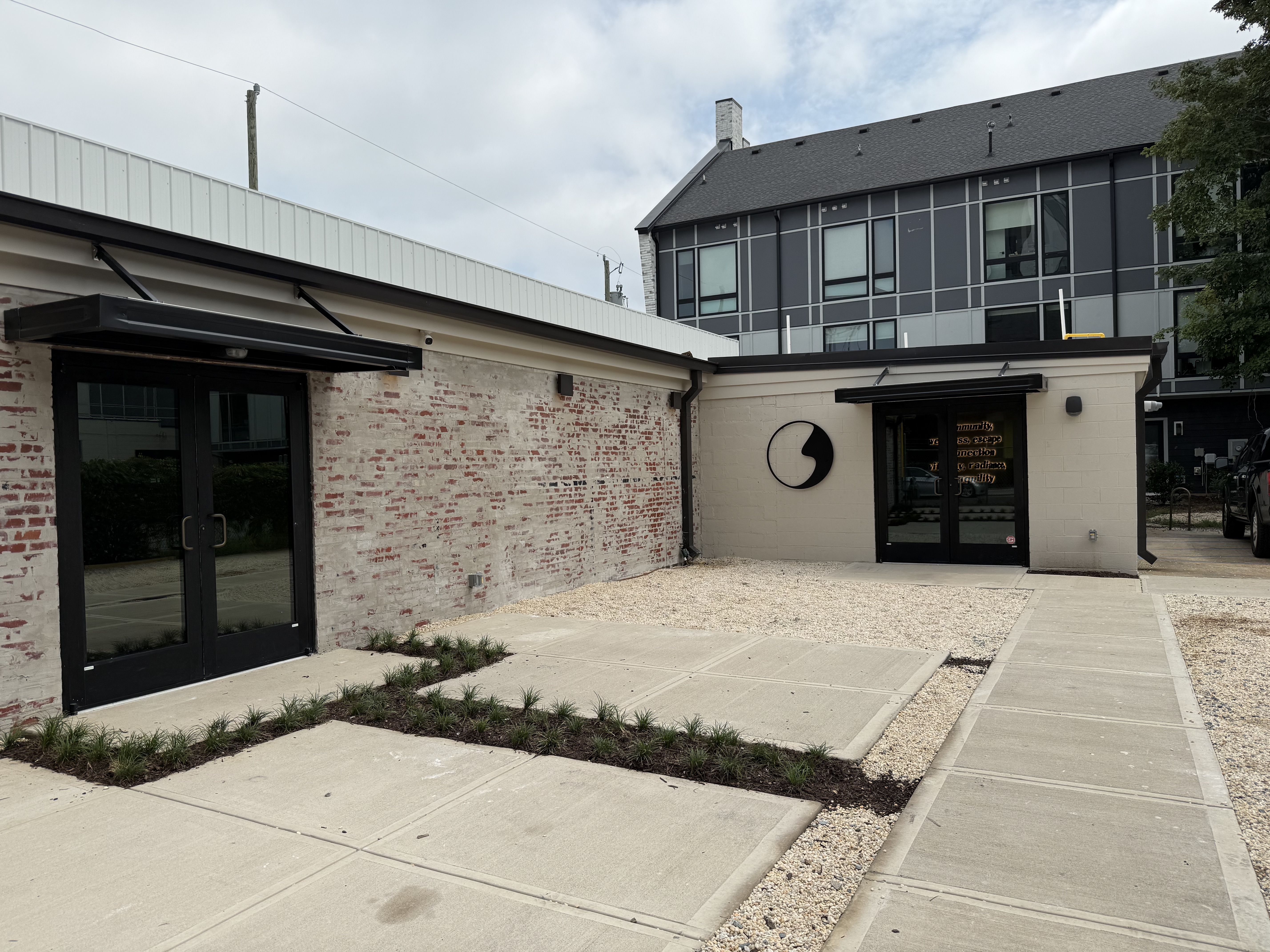 Outdoor area with concrete sidewalks, planted grass borders, a brick wall, and a building with black double doors and a circular black sign under cloudy sky.