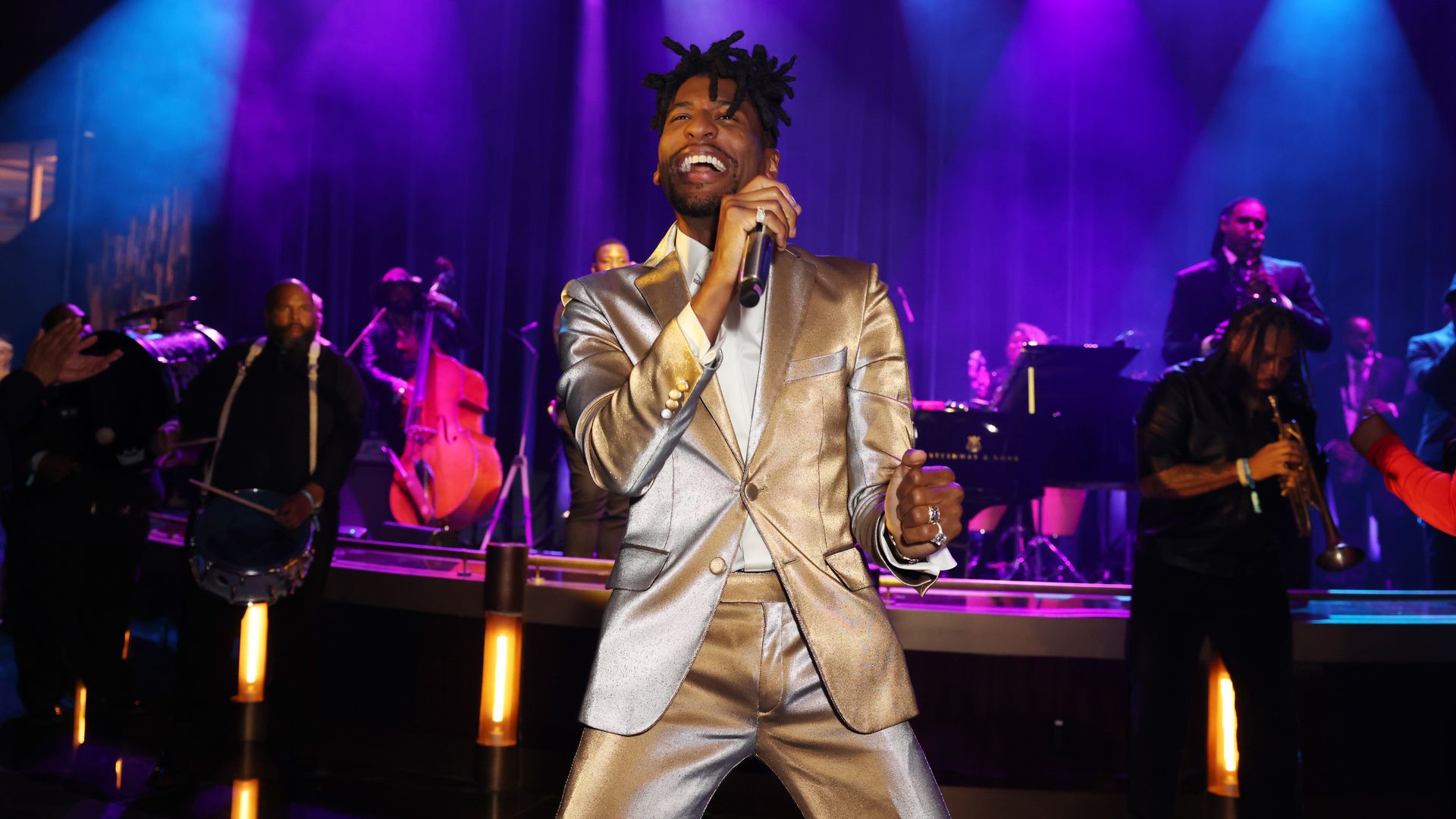 Jon Batiste, wearing a gold suit, sings into a microphone onstage.